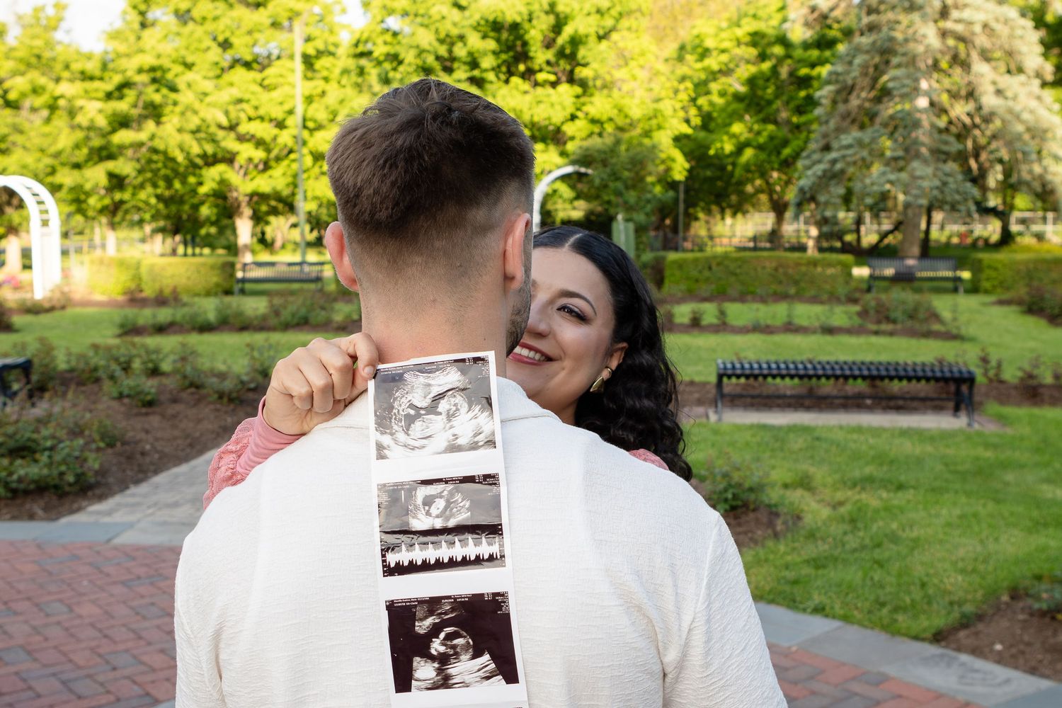 A smiling pregnant woman hugs her partner, who wears ultrasound photos draped over his shoulder, in a park.