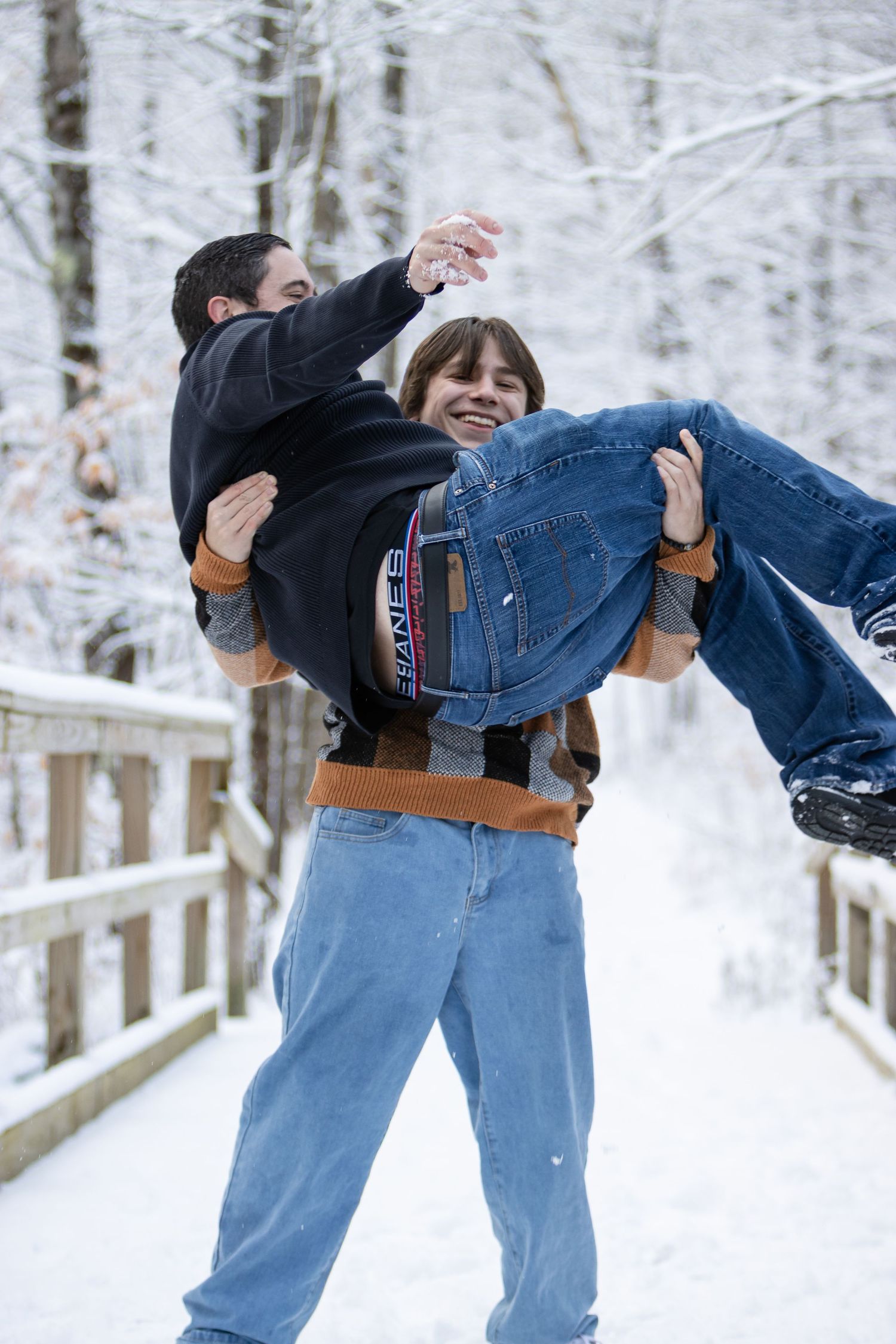 Teenage boy lifts a smiling person in his arms and playfully carries them on a snow-covered wooden bridge in a winter forest.