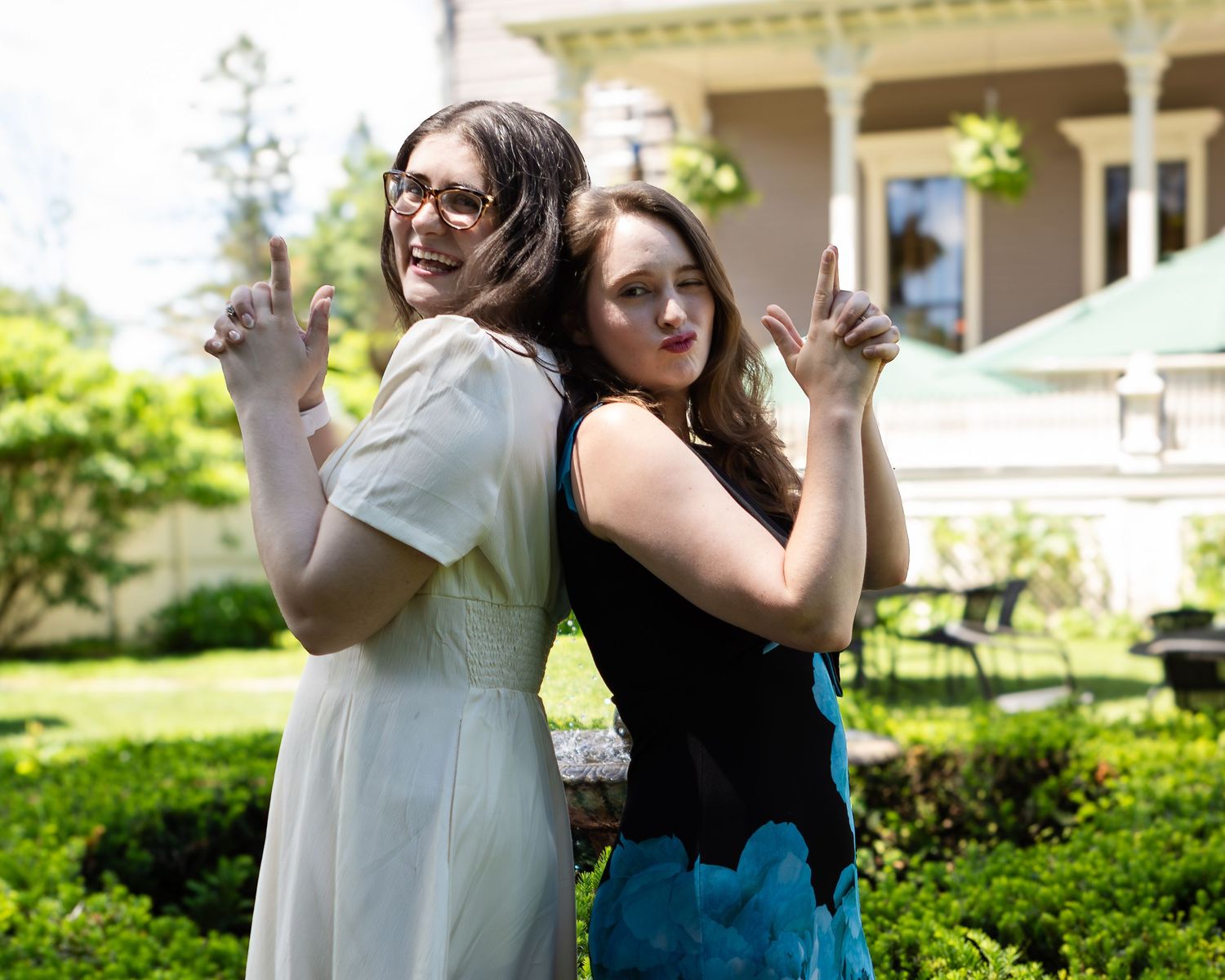 Two women stand back to back in a sunny garden, smiling and pointing finger guns, with a porch and green hedges behind them.