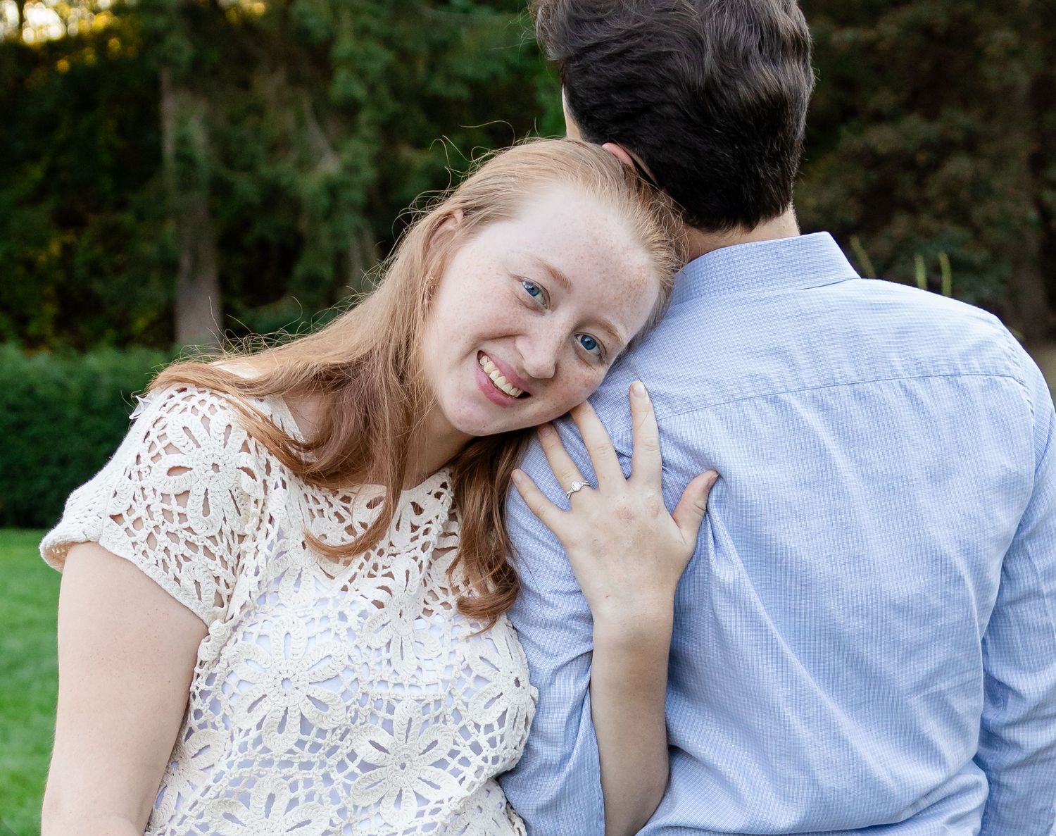 Smiling woman with long red hair rests her head against a man's shoulder in a green park during an outdoor couple portrait.