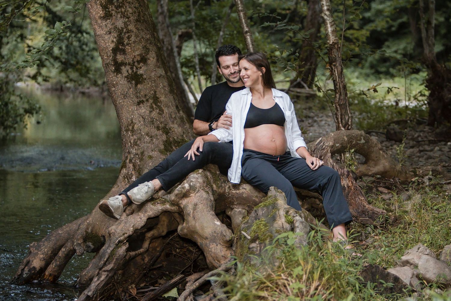 Expecting couple sitting together on a large tree root beside a calm river in a forest during a maternity photo session.