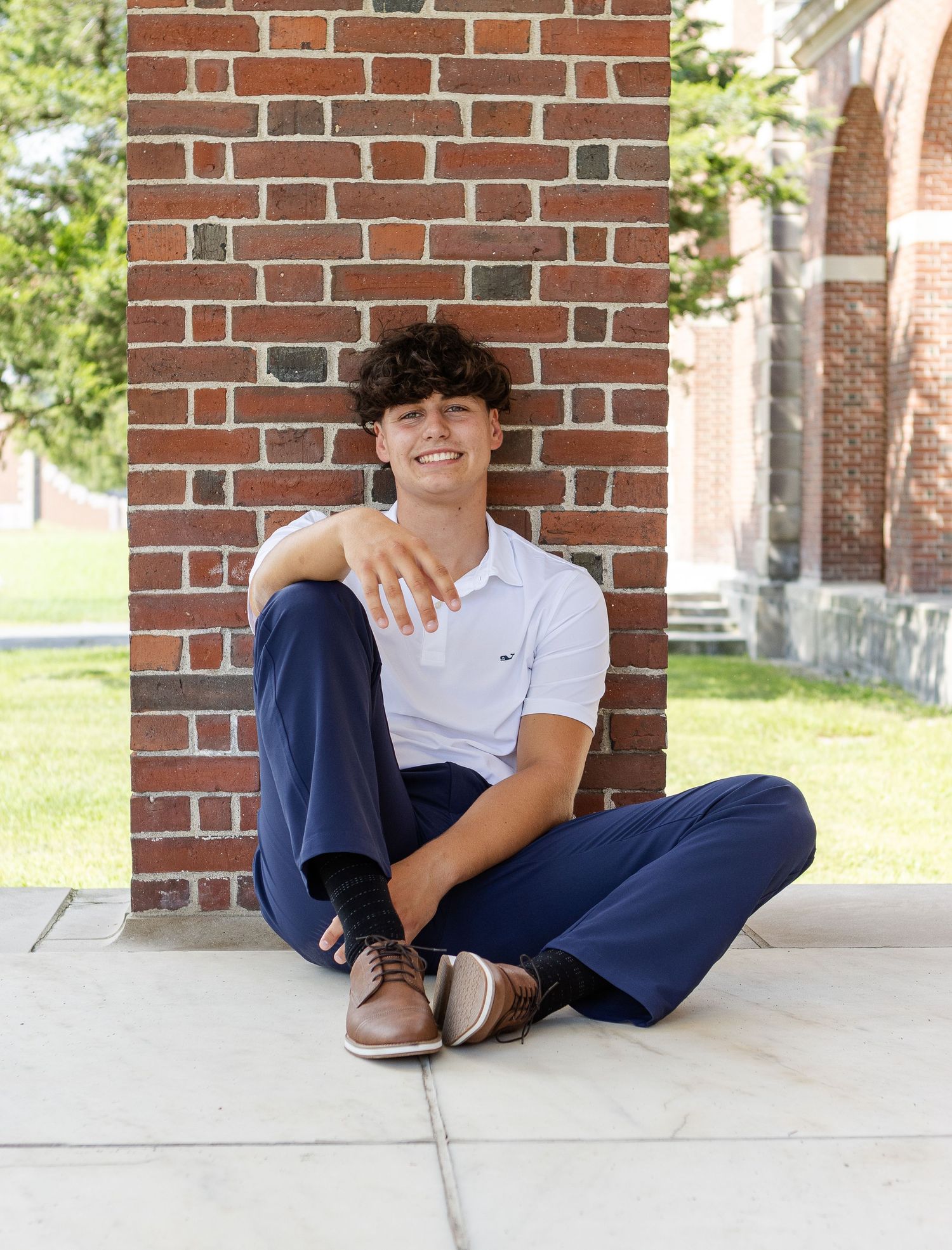 Smiling young man in a white polo and navy pants sits on a sidewalk leaning against a red brick pillar.