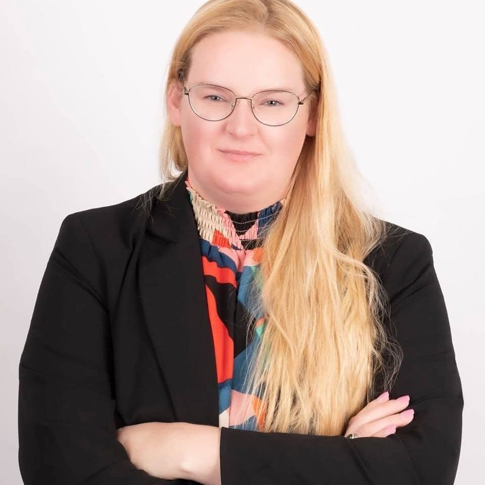 Professional headshot of a woman with long blonde hair and glasses, wearing a black blazer over a colorful top, standing with arms crossed.