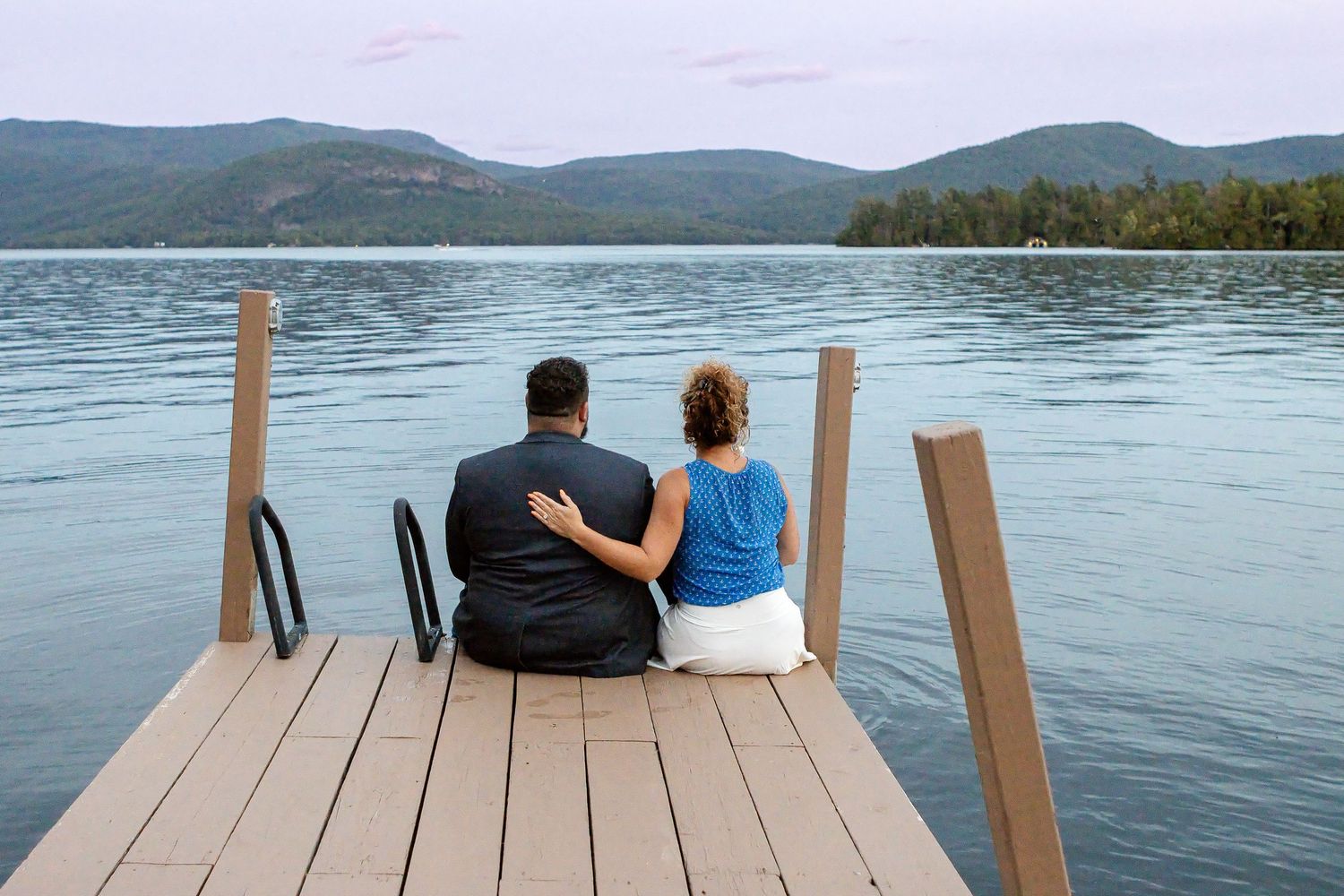 A newly engaged couple sits close together on the end of a wooden dock overlooking a calm lake and forested mountains.