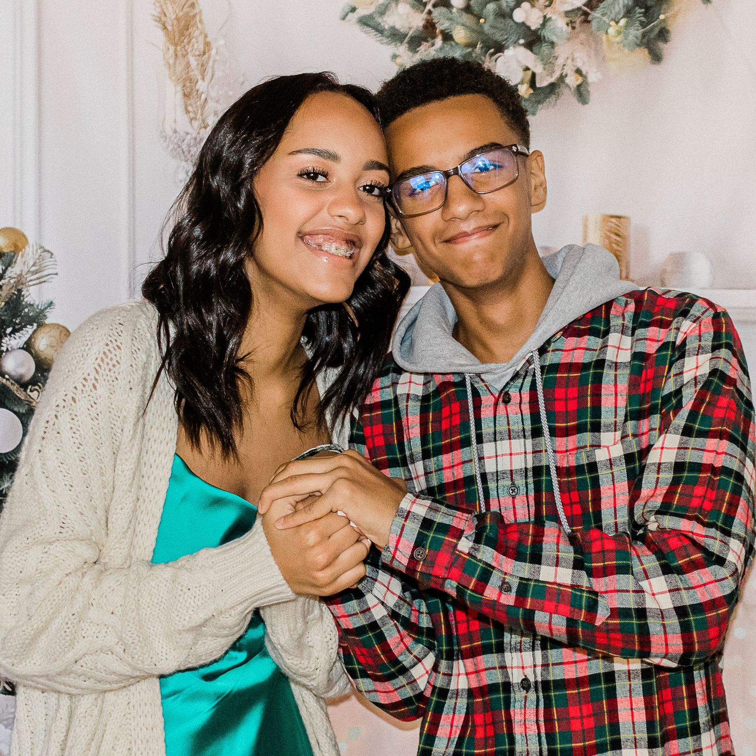 Smiling couple in cozy holiday outfits, a woman in teal satin and cardigan and a man in plaid hoodie, posing close by festive decor.