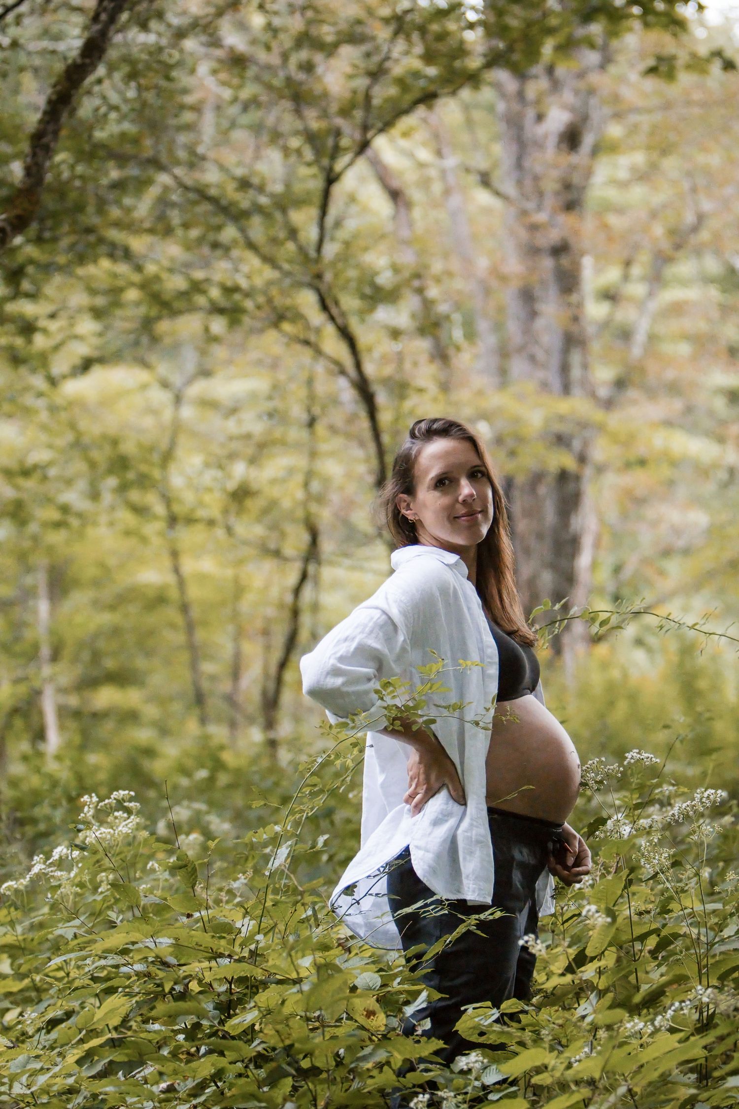 A smiling pregnant woman in a white button-up shirt and black top poses with a hand on her hip among leafy forest greenery.