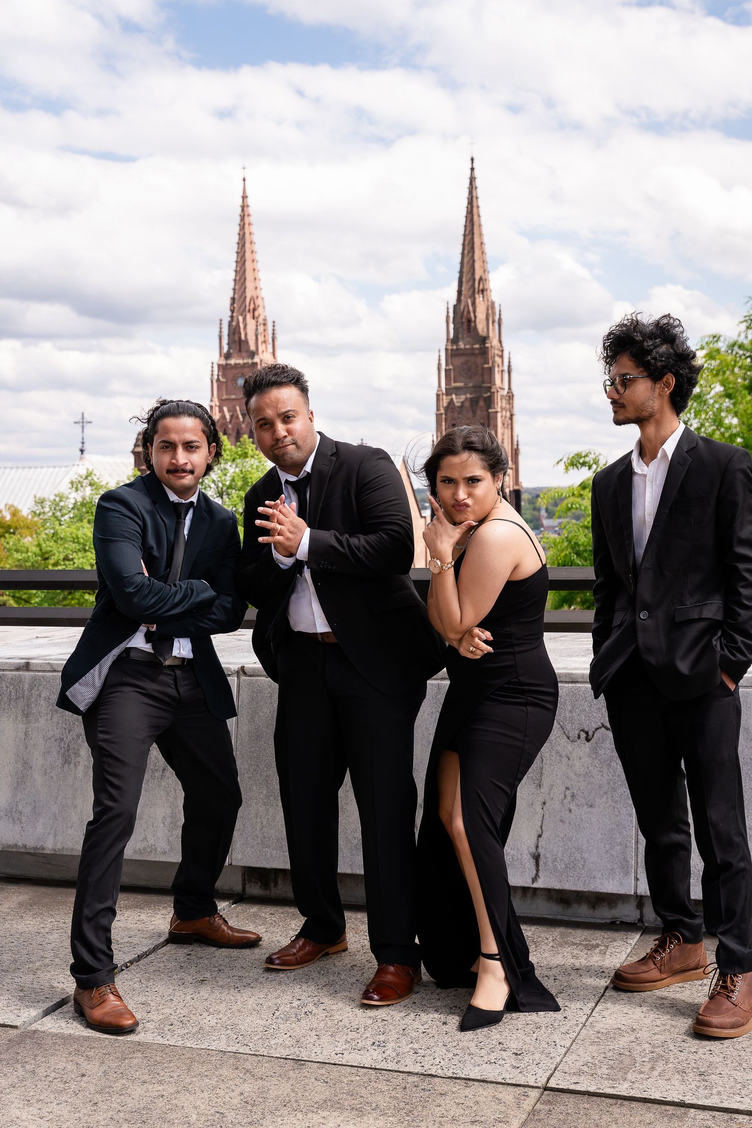 Four people in black formal outfits pose confidently on a rooftop terrace with Gothic church spires and a cloudy sky behind them.