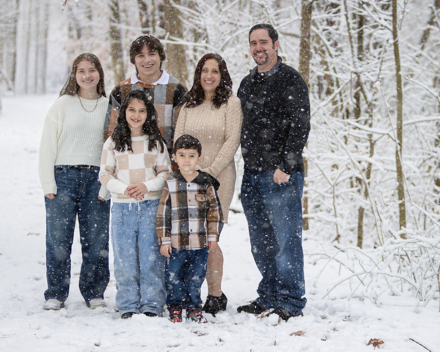 Family of five with two parents and three children poses together in a snowy forest as snowflakes fall around them.