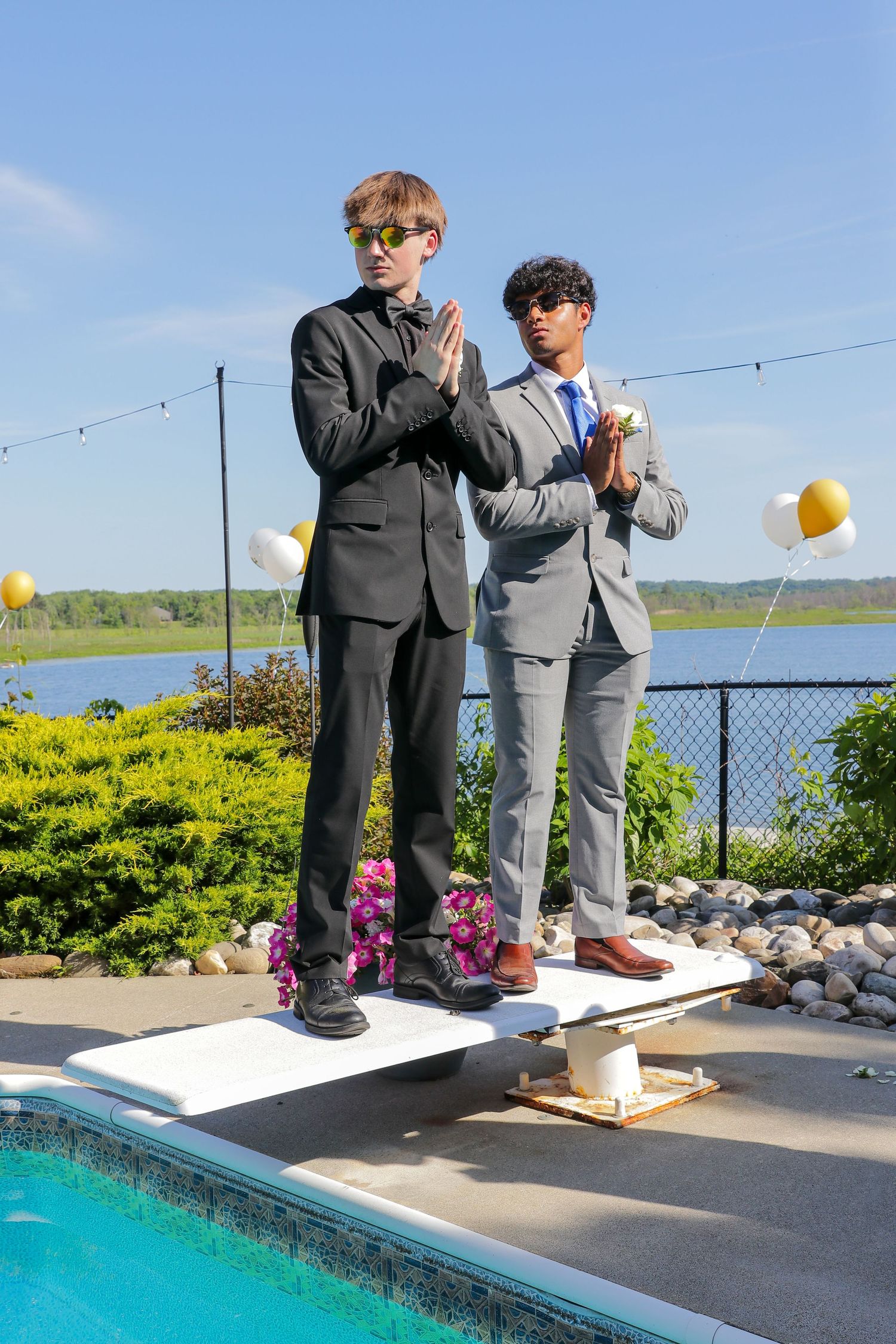 Two young men in formal suits and sunglasses stand on a diving board by a pool with balloons and a lake behind them.