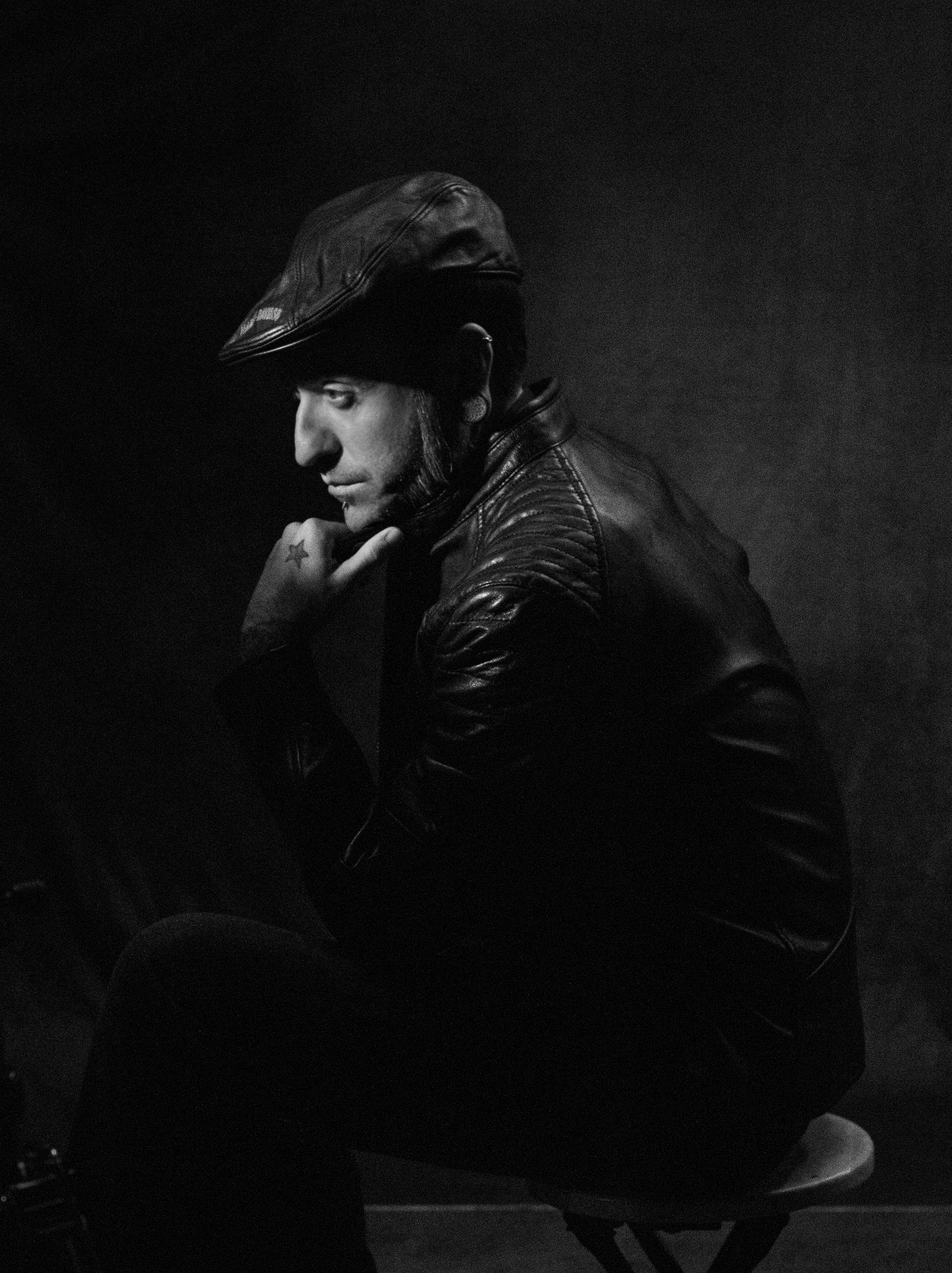 Black and white portrait of a pensive man in a leather jacket and flat cap seated on a stool with his hand at his chin.