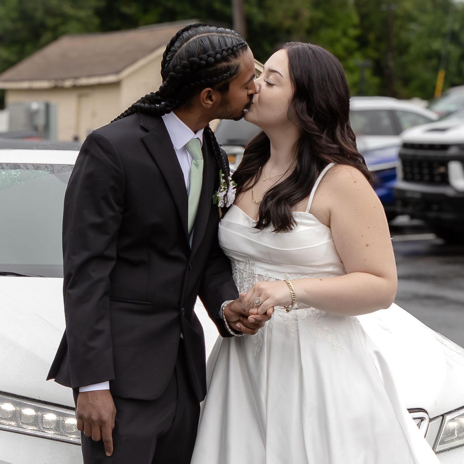 A bride in a white gown and a groom in a dark suit with braided hair share a kiss beside a white car.
