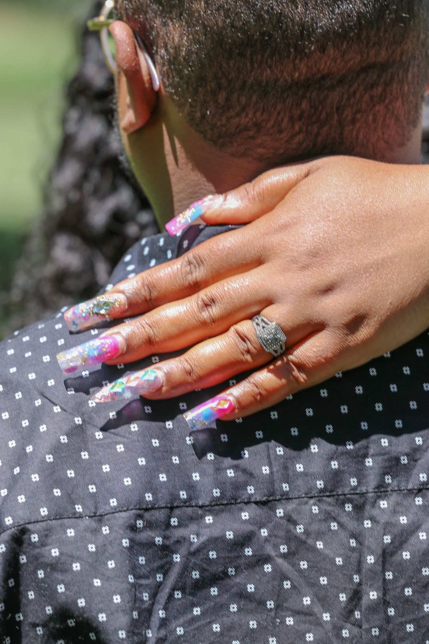 Close-up of a woman's hand with colorful painted nails and a ring resting on the neck of a man in a polka dot shirt.