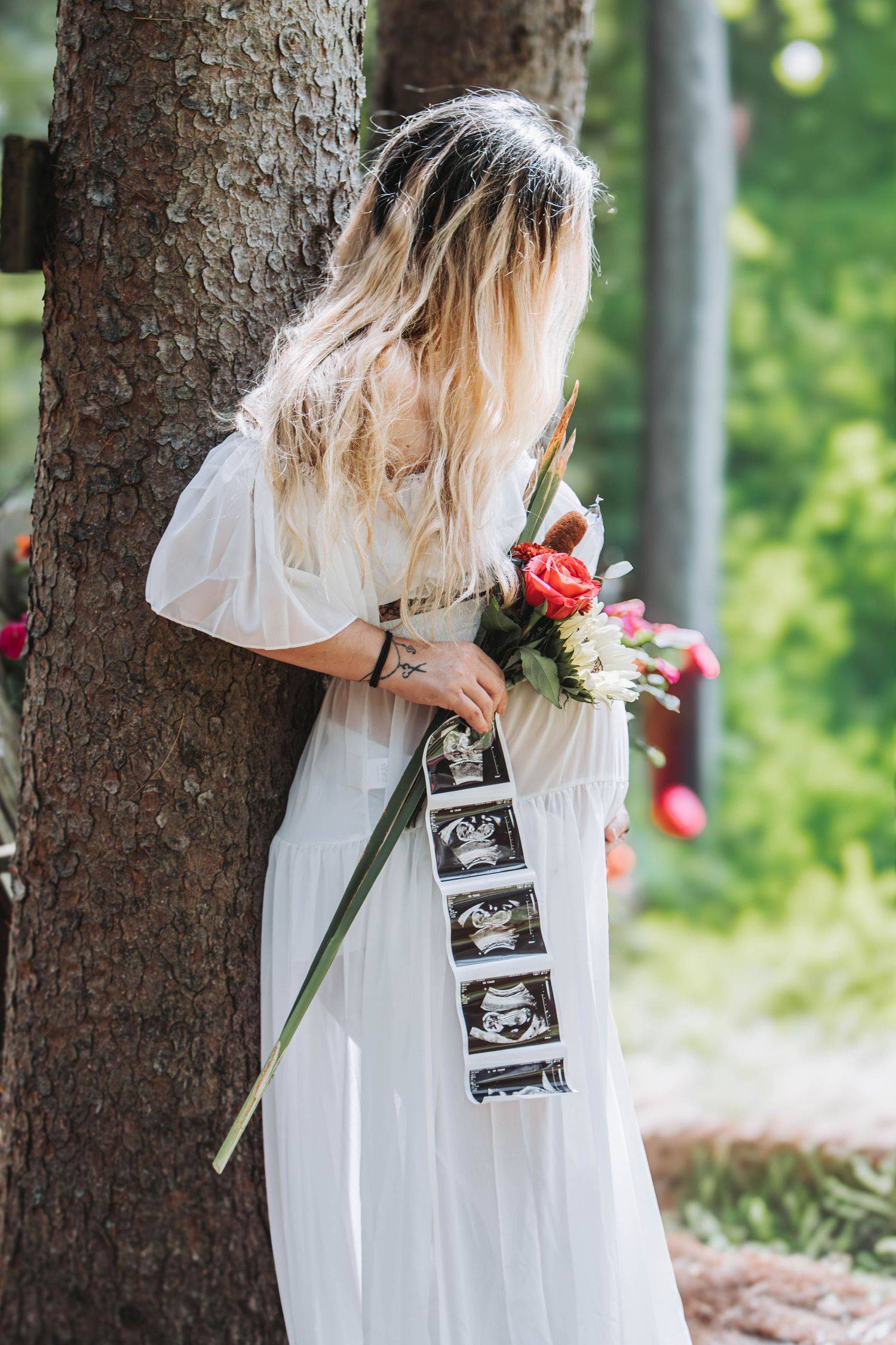 Woman in a white dress leans against a tree holding a bouquet and a strip of ultrasound photos.