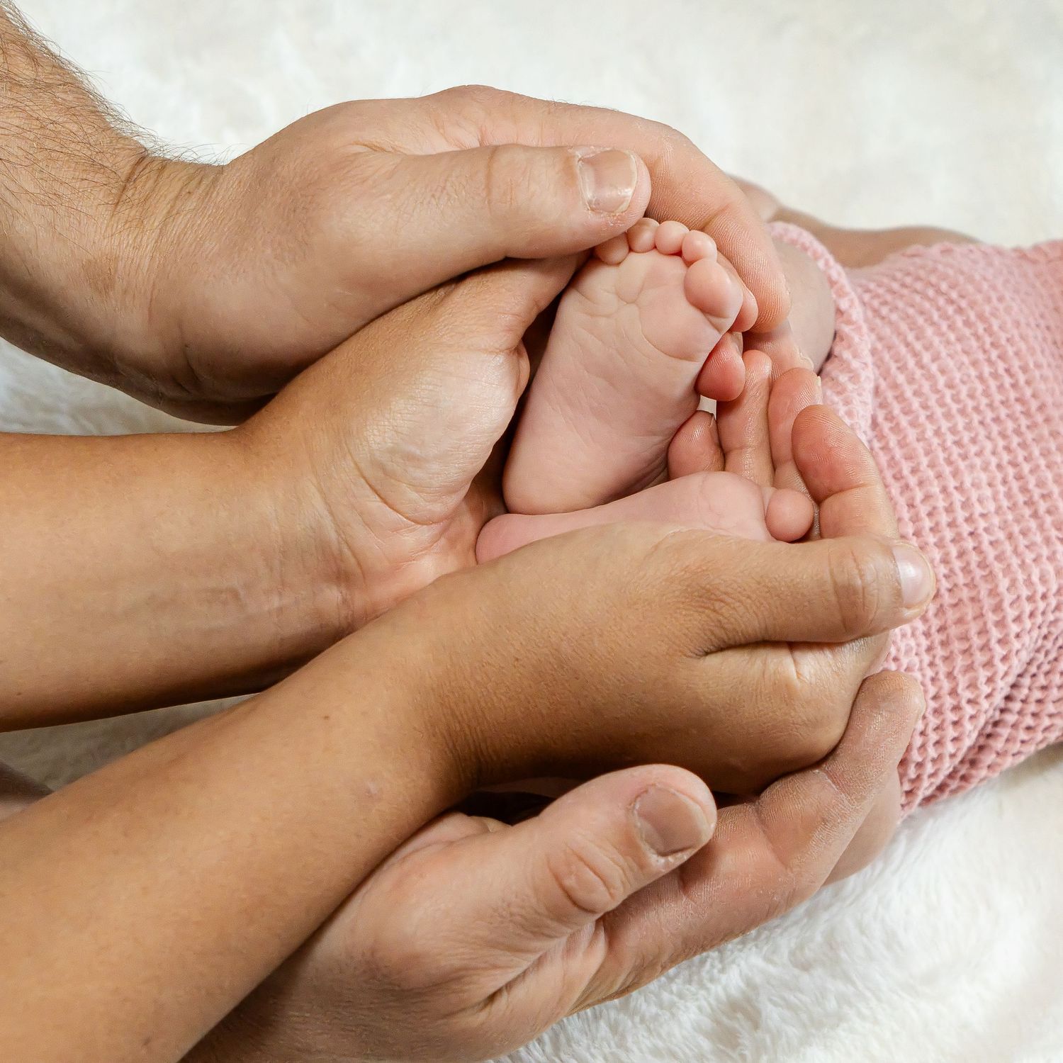 Parents' hands gently cradle a newborn baby's tiny feet on a soft white blanket next to a pink knit garment.