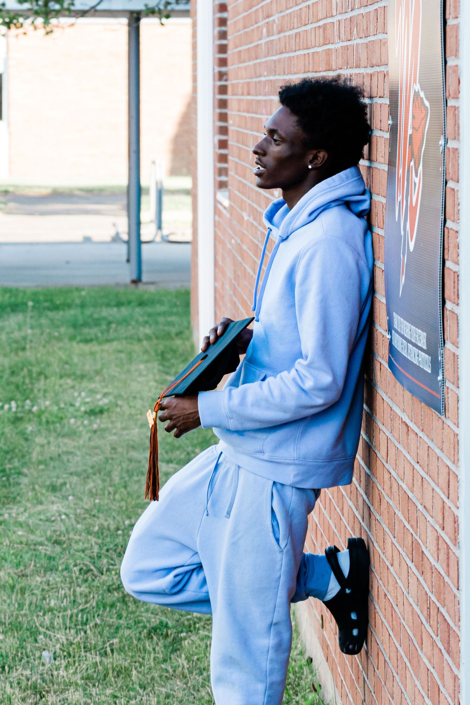 Young man in a light blue sweatsuit leans against a brick school wall holding a graduation cap with a tassel.
