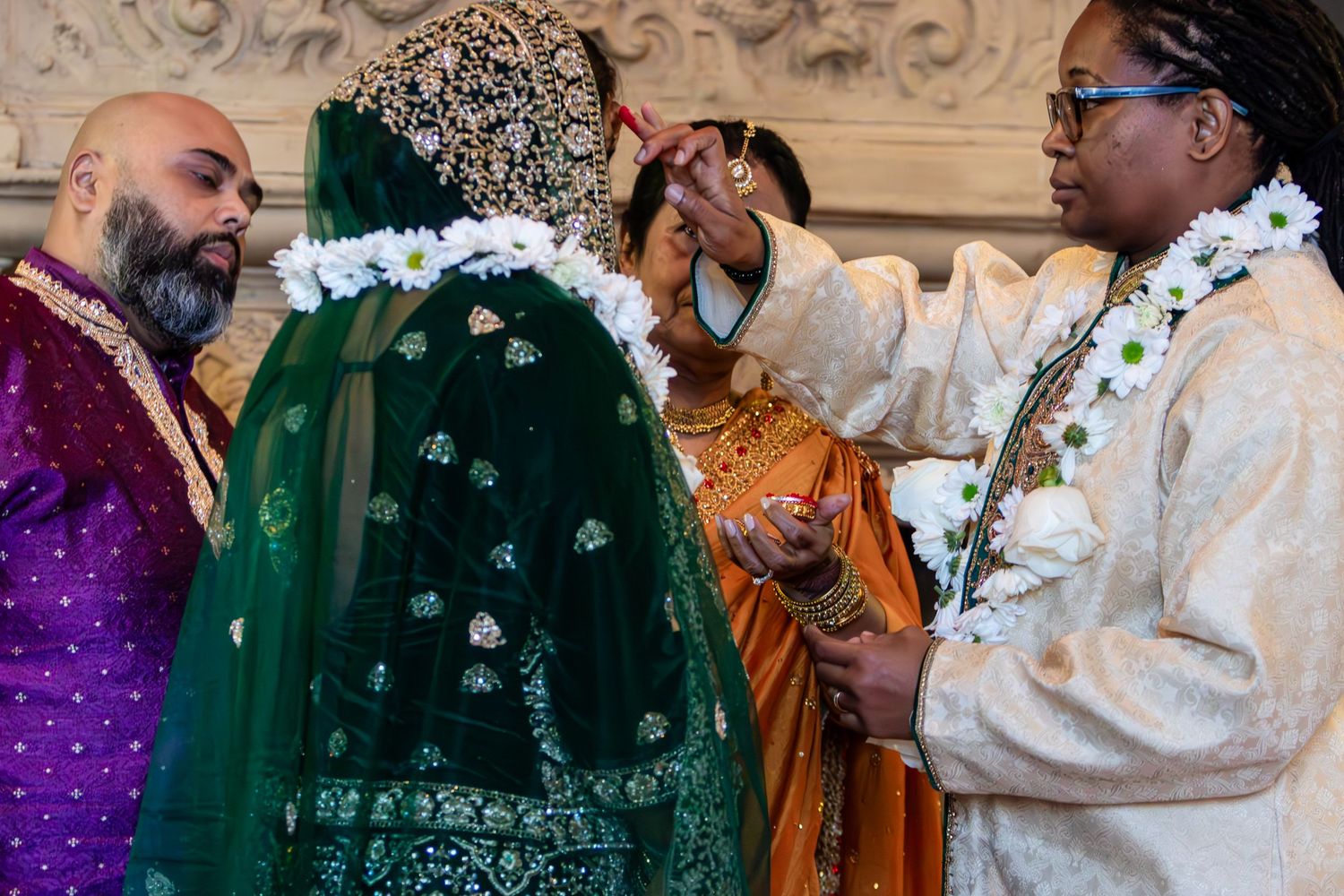 A bride in an embroidered green veil receives a floral garland during a wedding ceremony as a groom and an officiant look on.