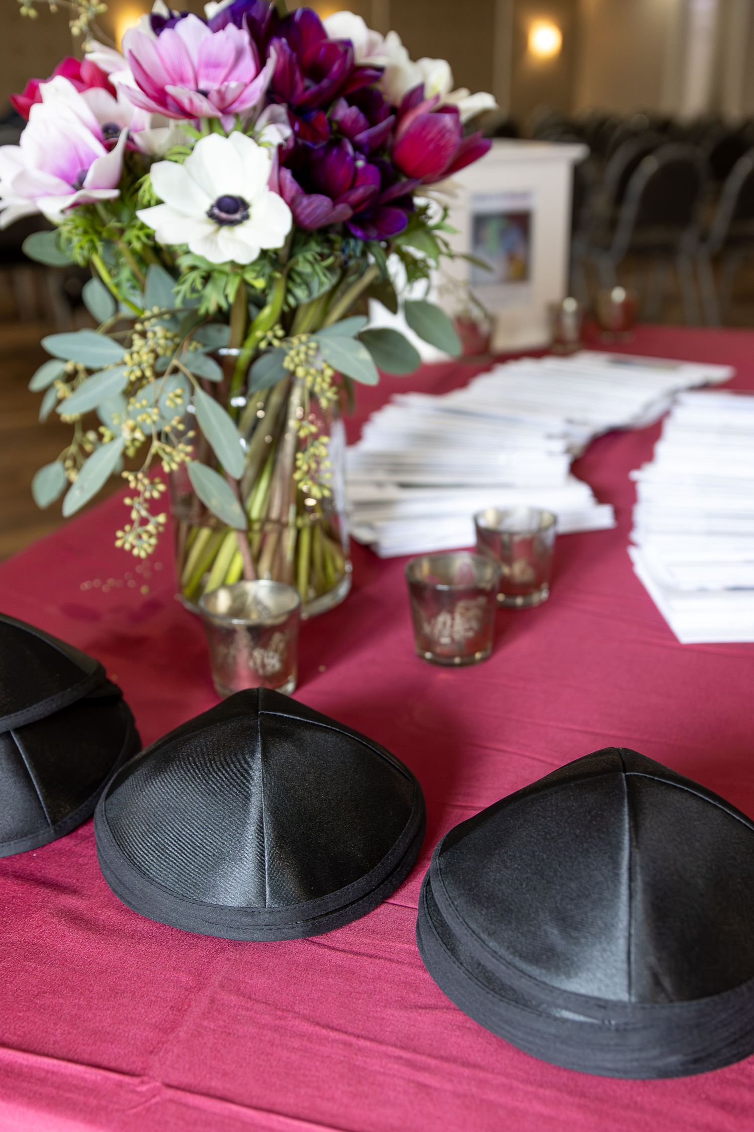 Black satin yarmulkes on a burgundy tablecloth beside a floral arrangement of anemones, votive holders and stacks of programs at an event.