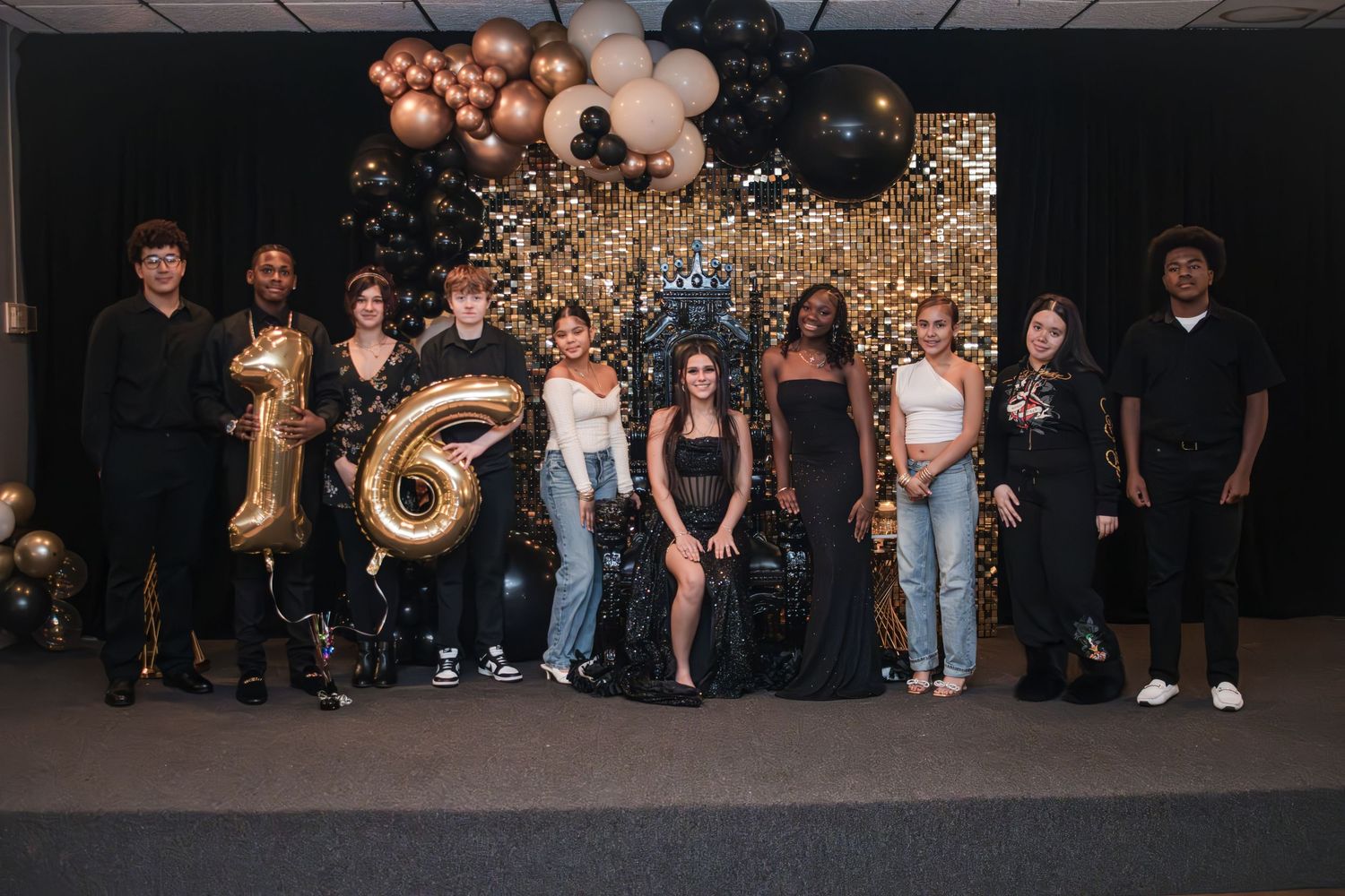Group of ten teens in black and white outfits posing on stage with gold number 16 balloons and a black, gold and white balloon backdrop.