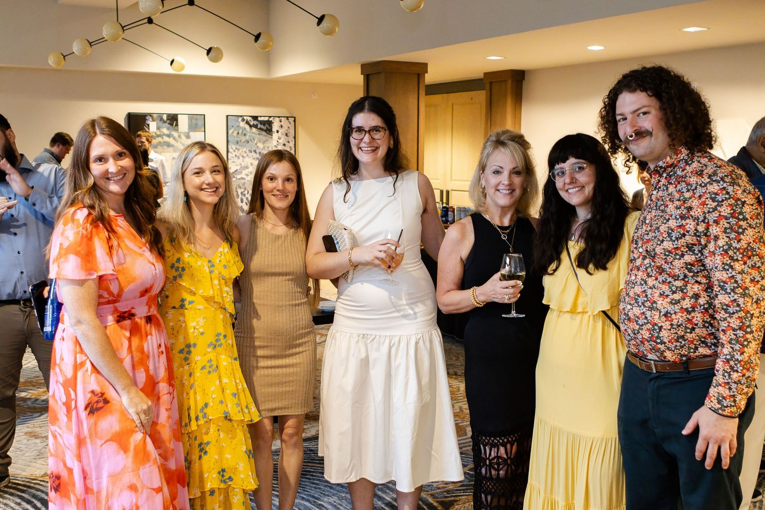 Group of smiling men and women in colorful dresses and patterned shirts pose together at an indoor social gathering with drinks.