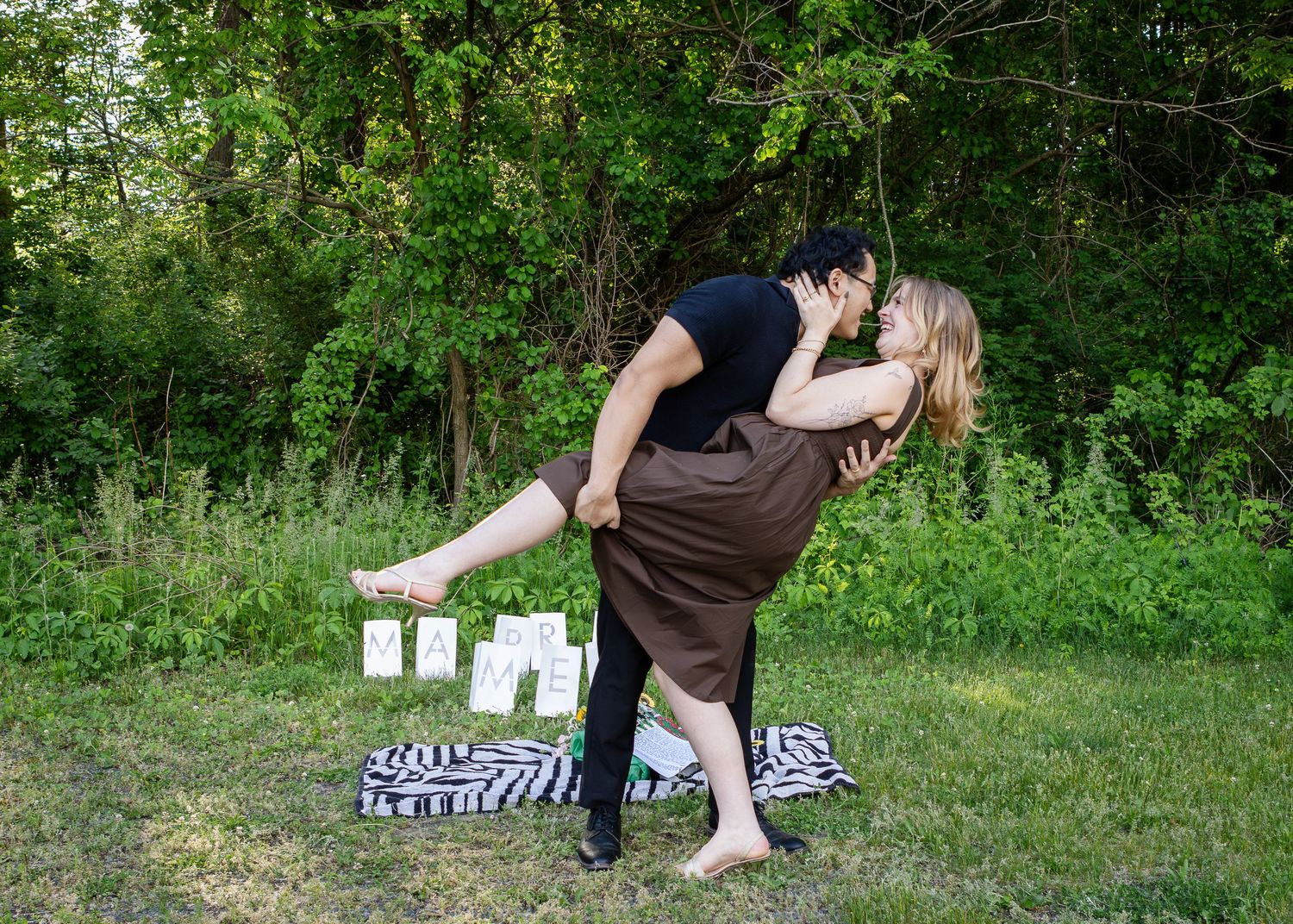 Couple sharing a romantic dip kiss on a grassy lawn surrounded by trees, with a striped blanket and small signs behind them.