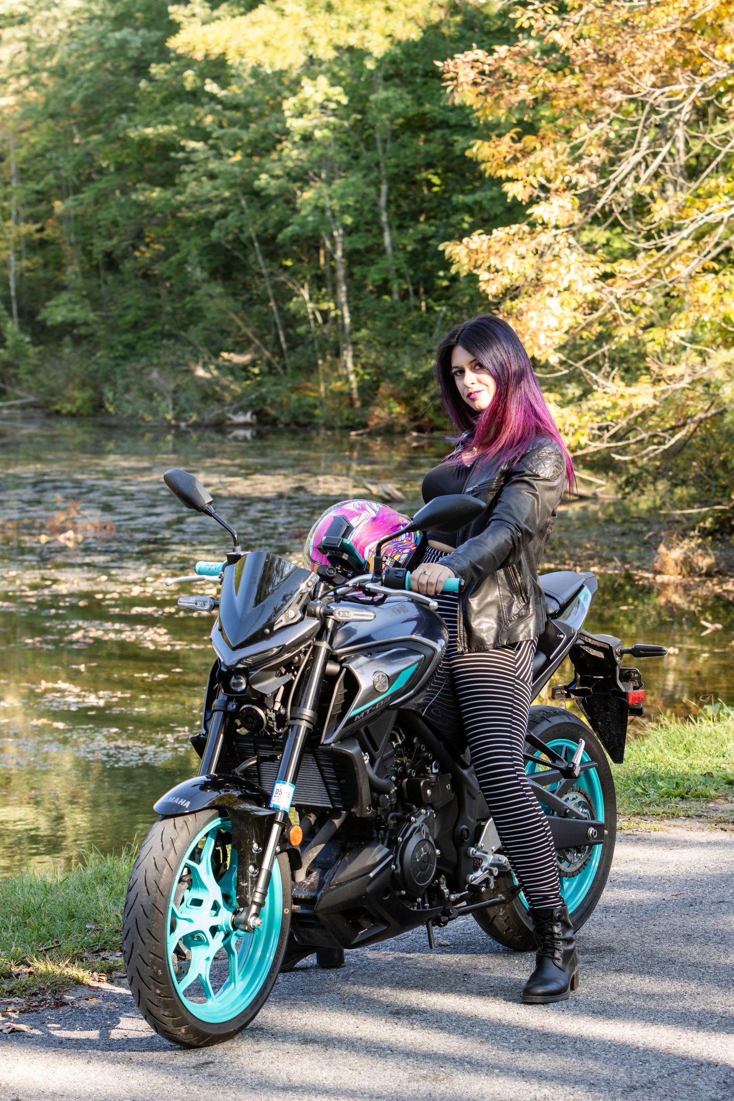 Woman with purple hair and striped leggings sits on a black motorcycle with teal wheels beside a pond on a sunny autumn day.