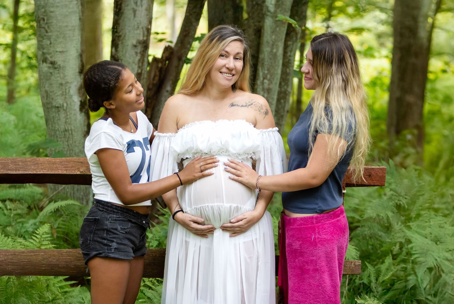 Two women place their hands on a pregnant woman's belly in a white dress during an outdoor forest maternity photo.