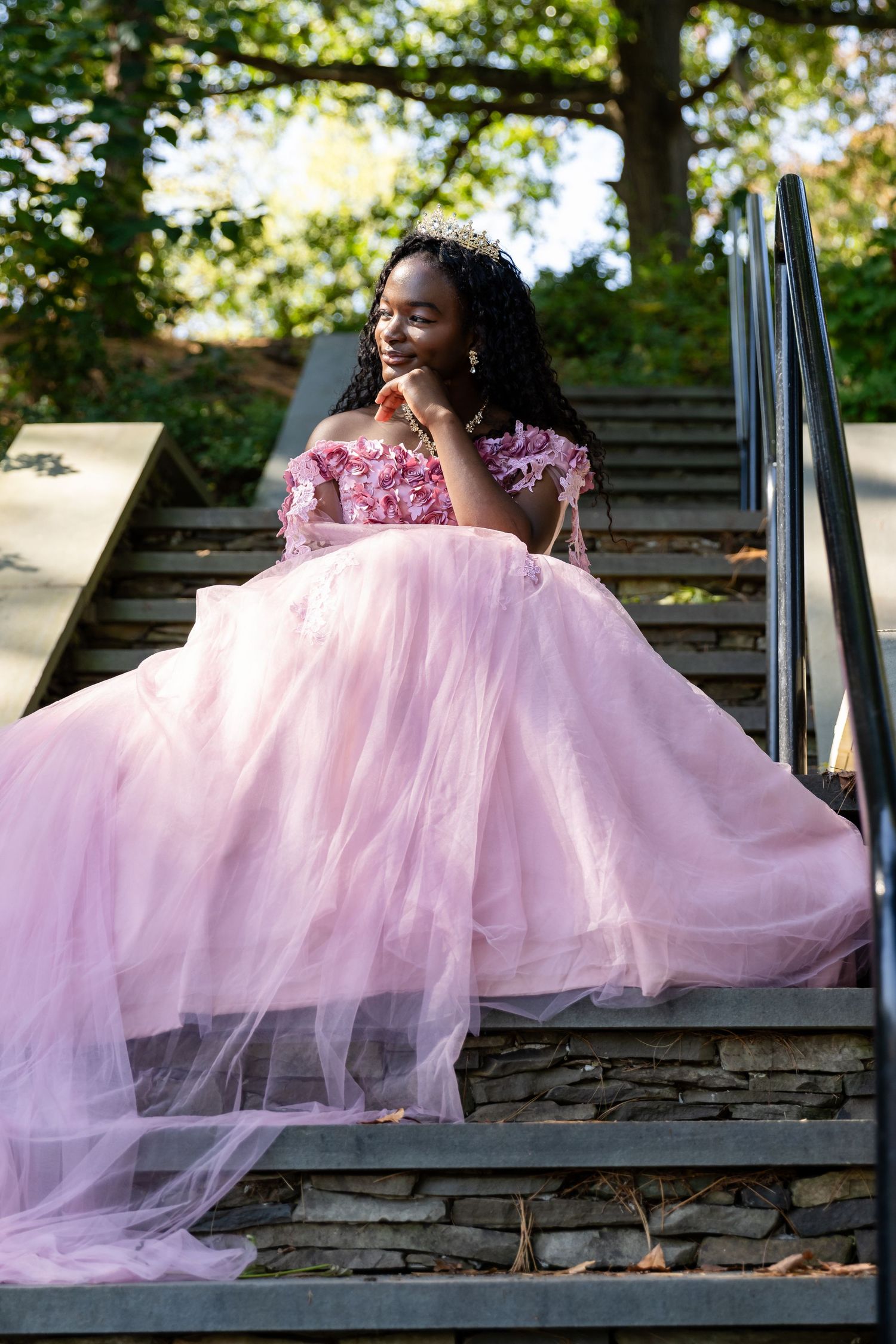 Young woman in a flowing pink tulle gown and tiara sits on stone steps with her hand near her face.
