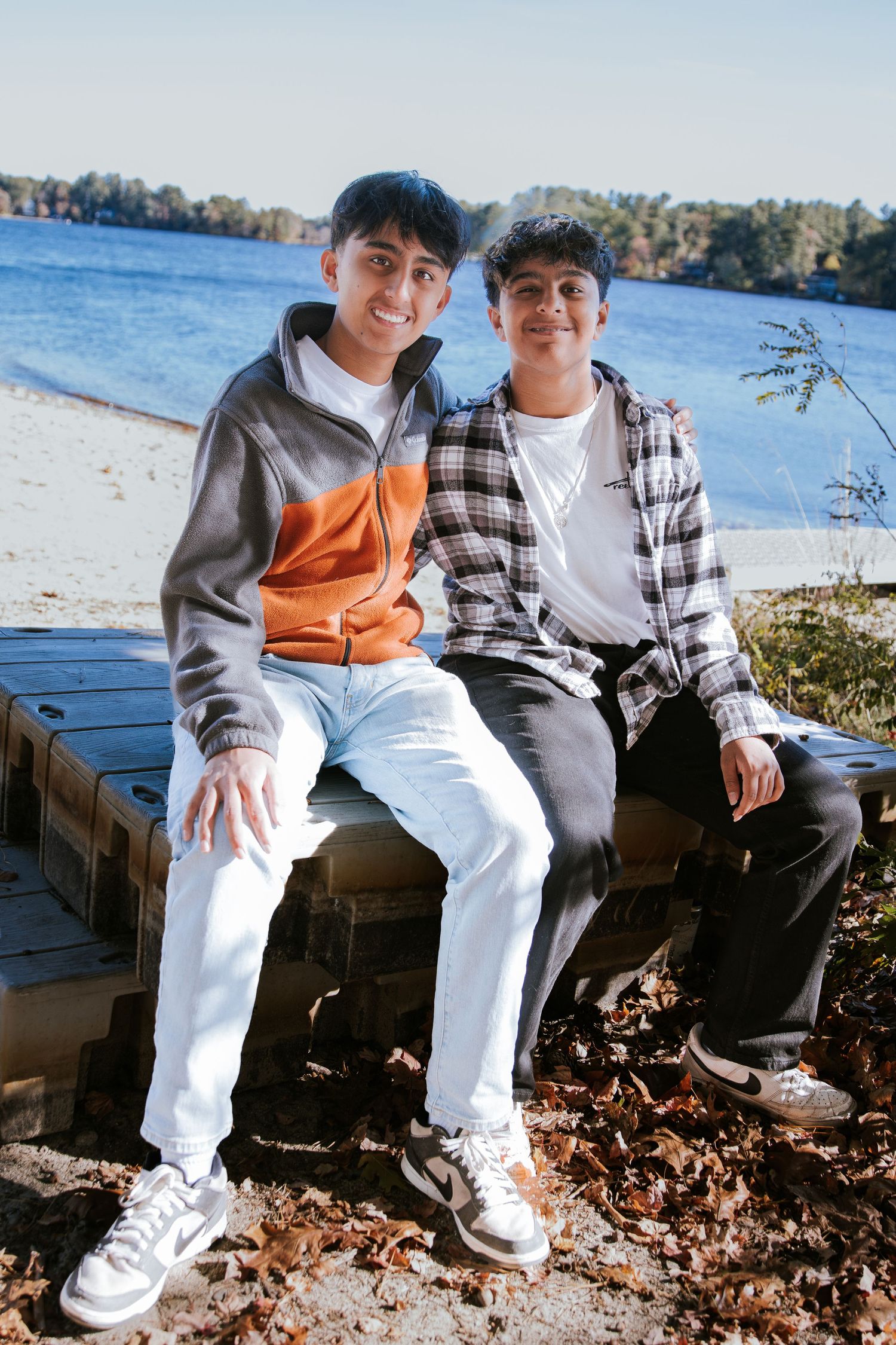 Two smiling teenage boys sit side by side on a wooden dock by a lake, surrounded by fallen autumn leaves.