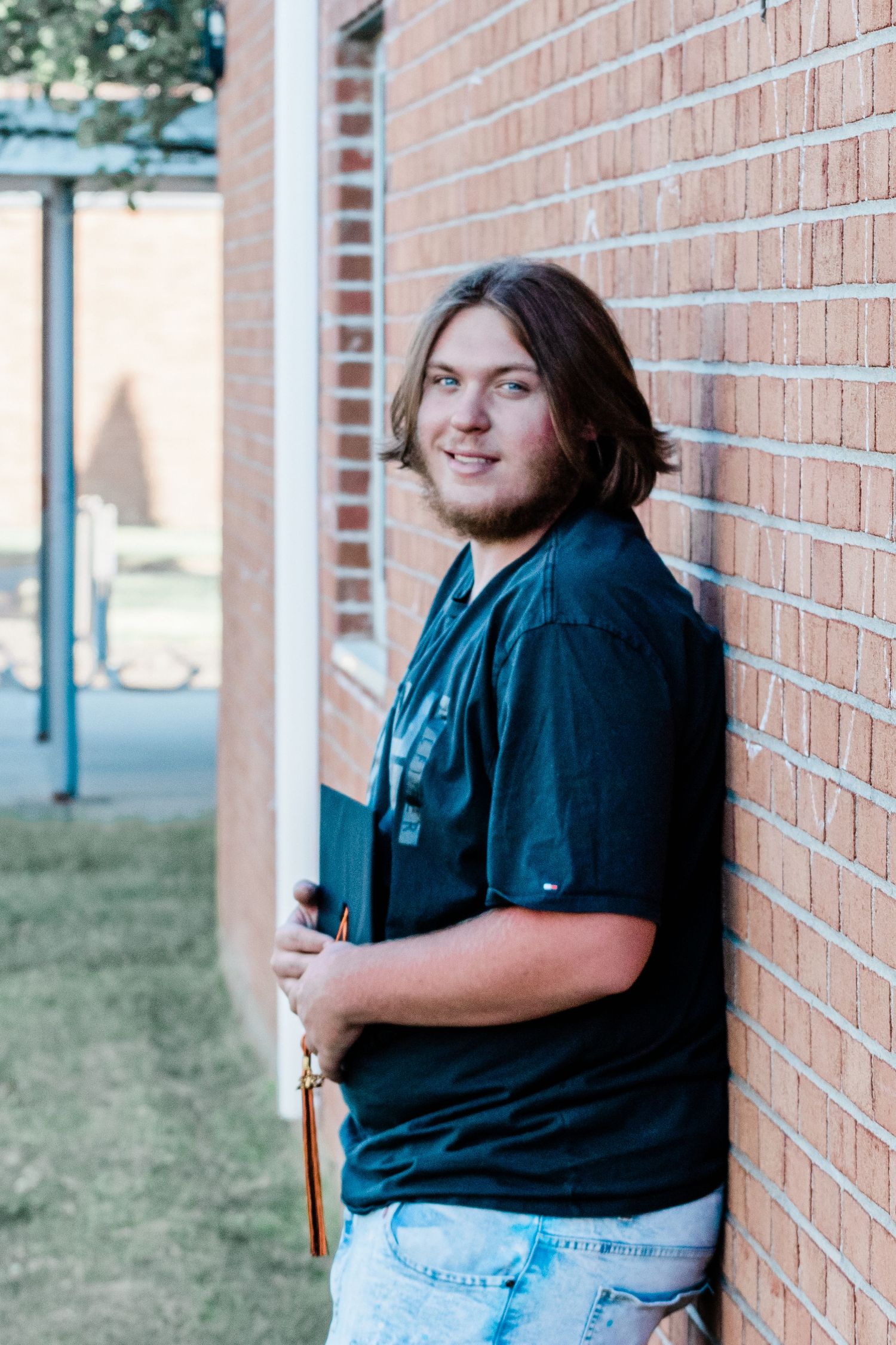 Smiling young man with shoulder-length hair and a beard leans against a brick wall holding a graduation cap with a tassel.