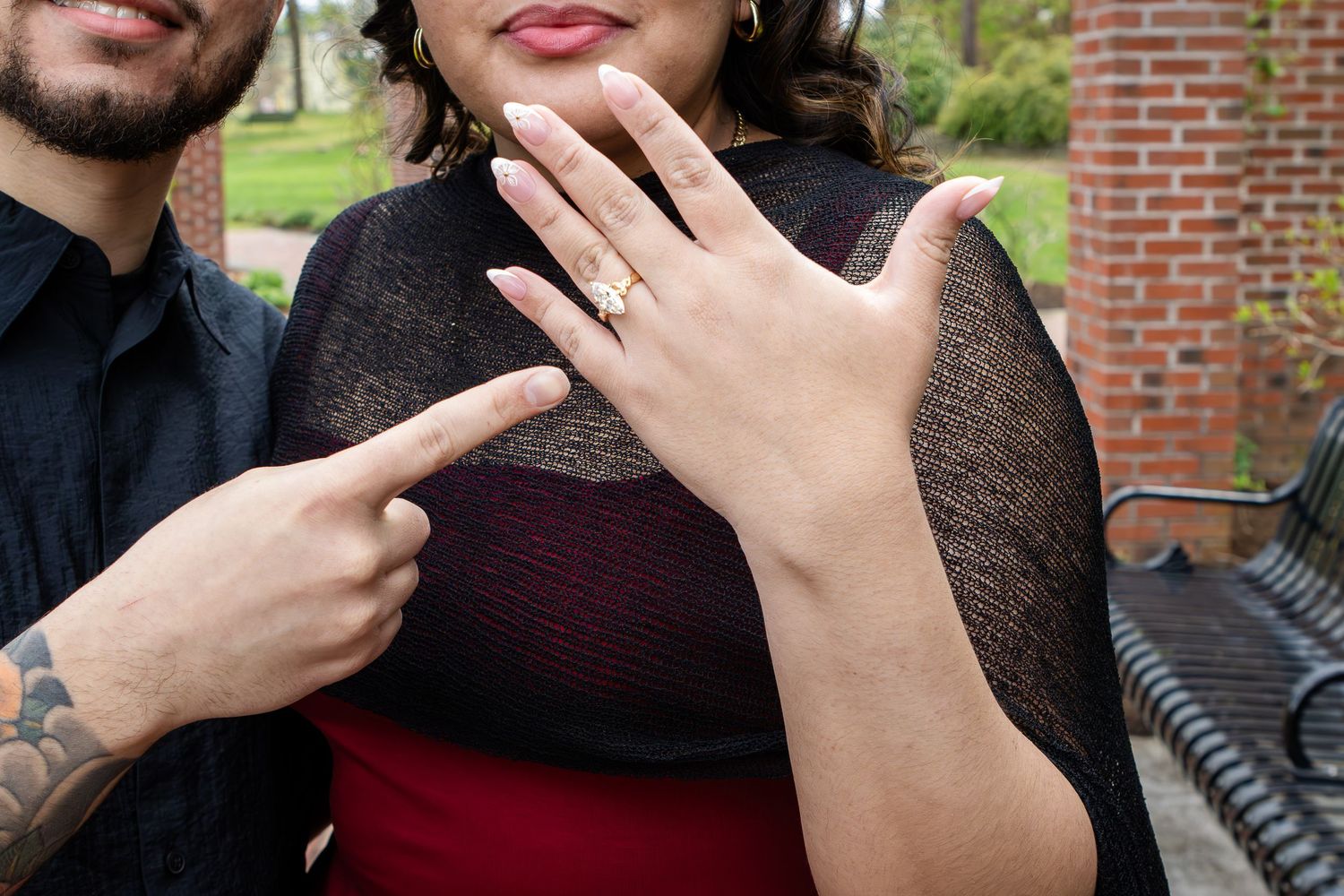 Engaged couple outdoors as the woman shows her diamond engagement ring while the man points to it, with a brick pillar behind them.