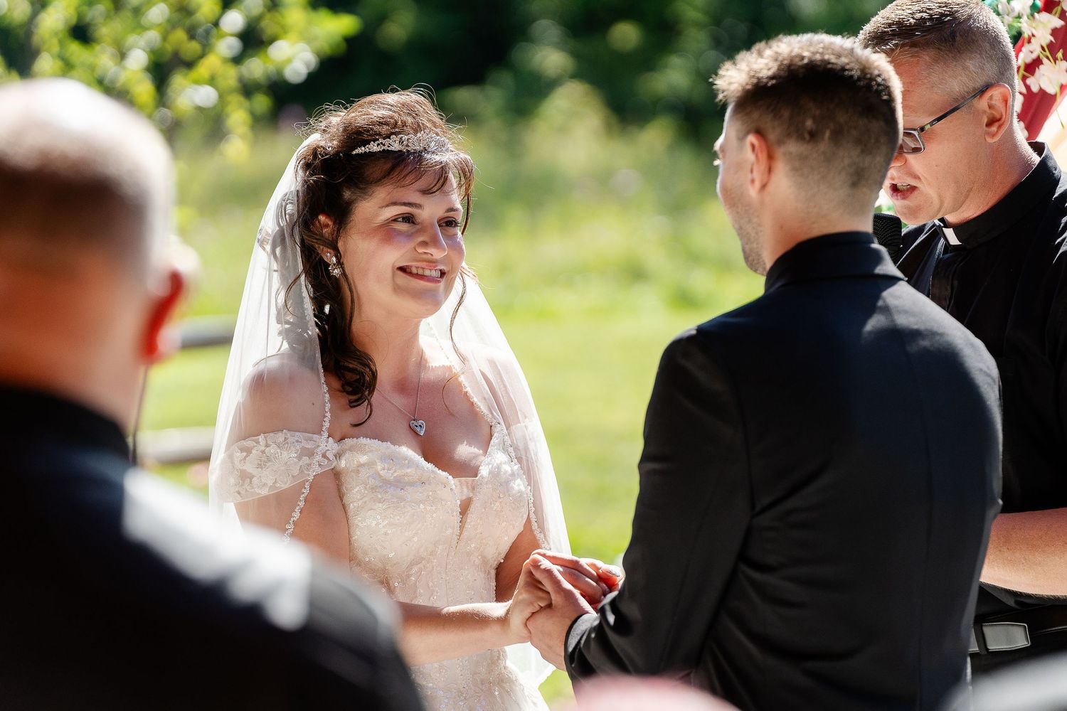 A smiling bride in a lace gown and veil holds hands with her groom during an outdoor wedding ceremony as the officiant looks on.