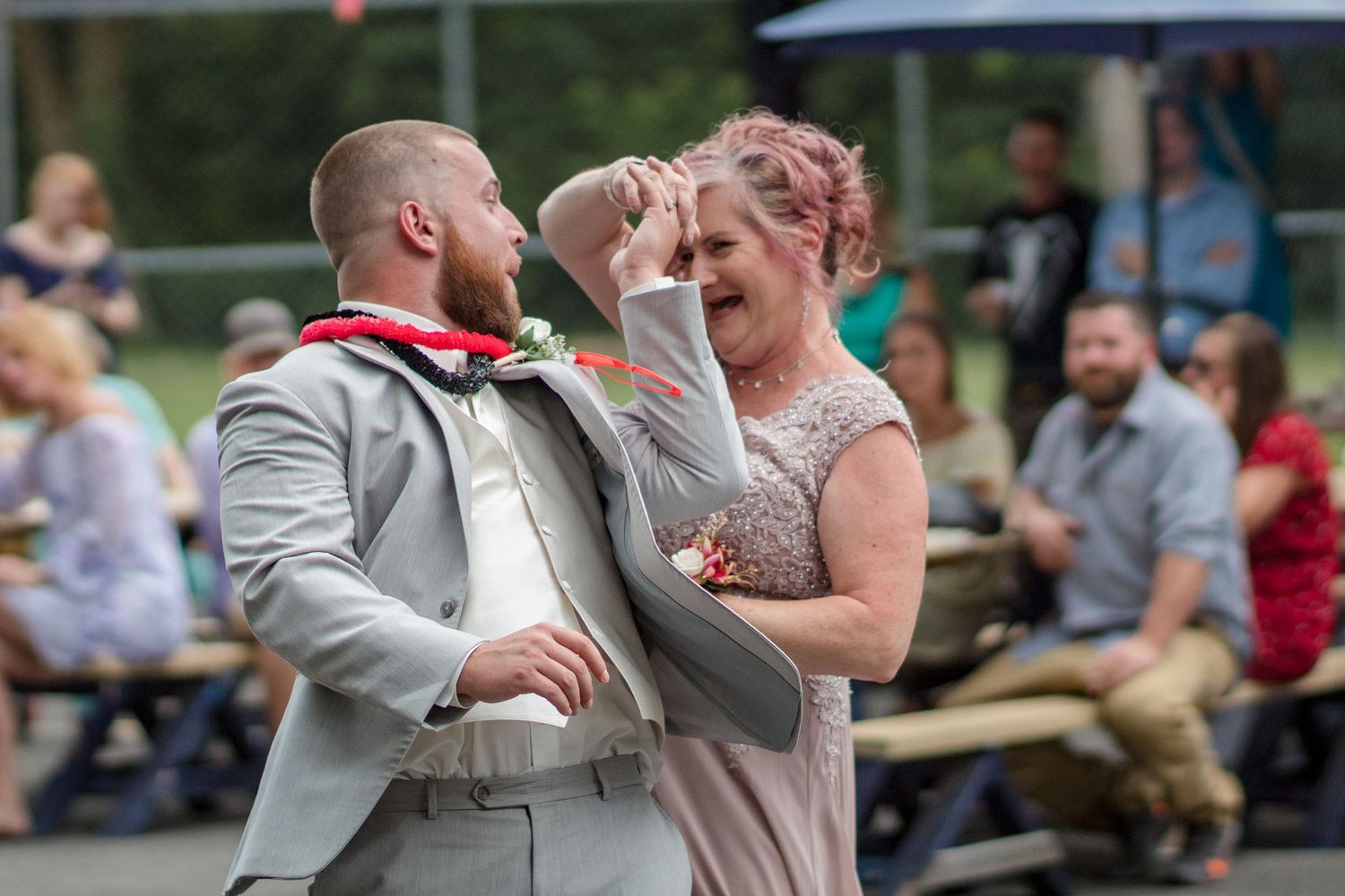 A groom in a gray suit and a woman in a floral dress dance joyfully at an outdoor wedding reception as guests watch.