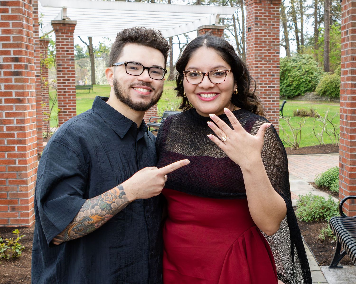 A smiling couple in glasses stands in a brick garden pavilion as the woman shows off her new engagement ring.