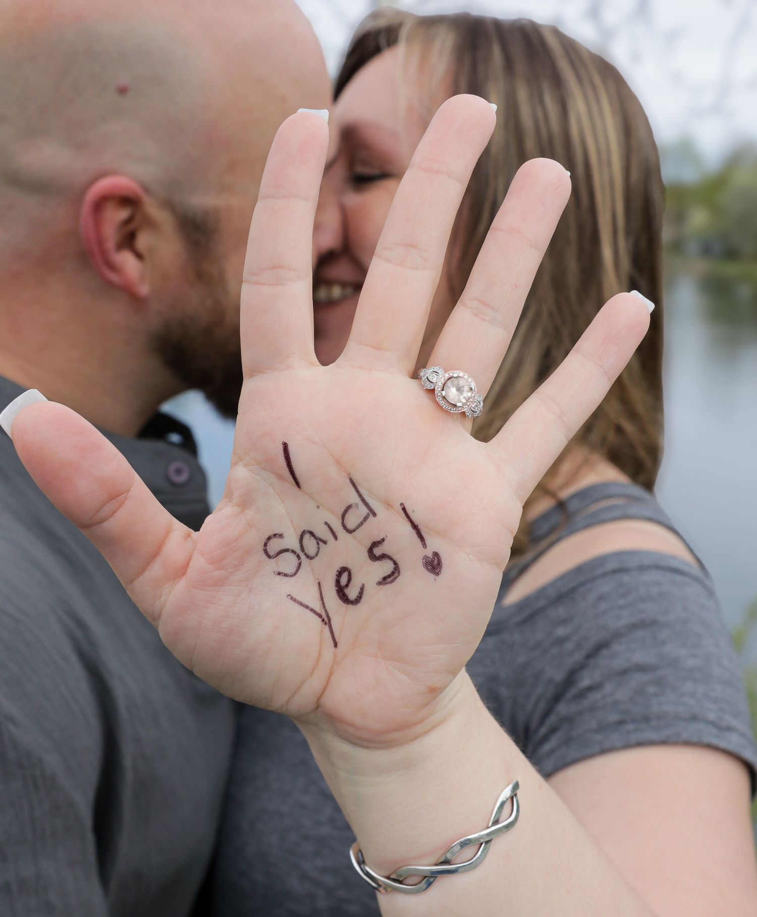 A woman's hand with a sparkling engagement ring shows the handwritten words I said yes while a couple embraces behind it.