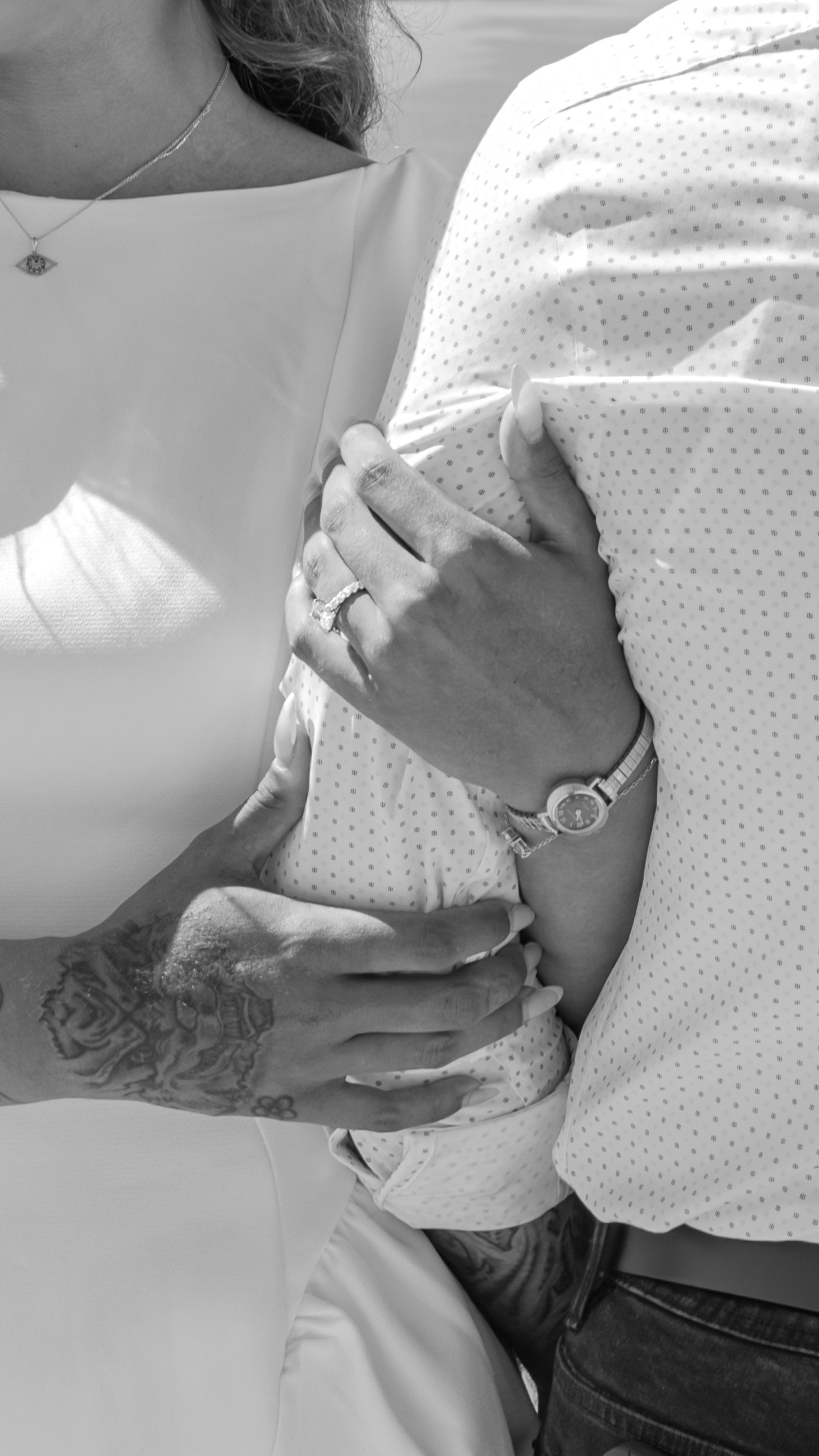 Black and white close-up of a couple linking arms, with a woman's hand wearing a diamond ring resting on the man's patterned shirt sleeve.