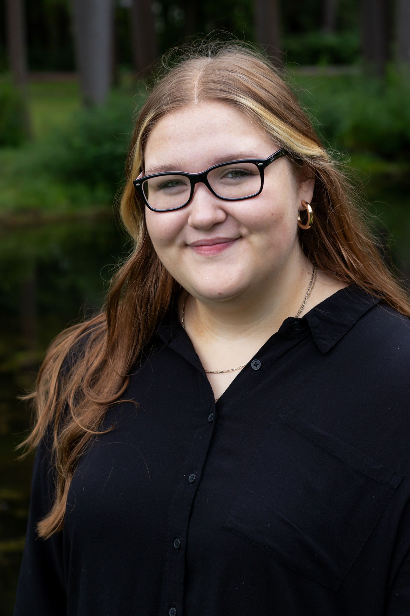 Portrait of a smiling young woman with glasses, long reddish-blonde hair and gold hoop earrings in a black shirt outdoors.