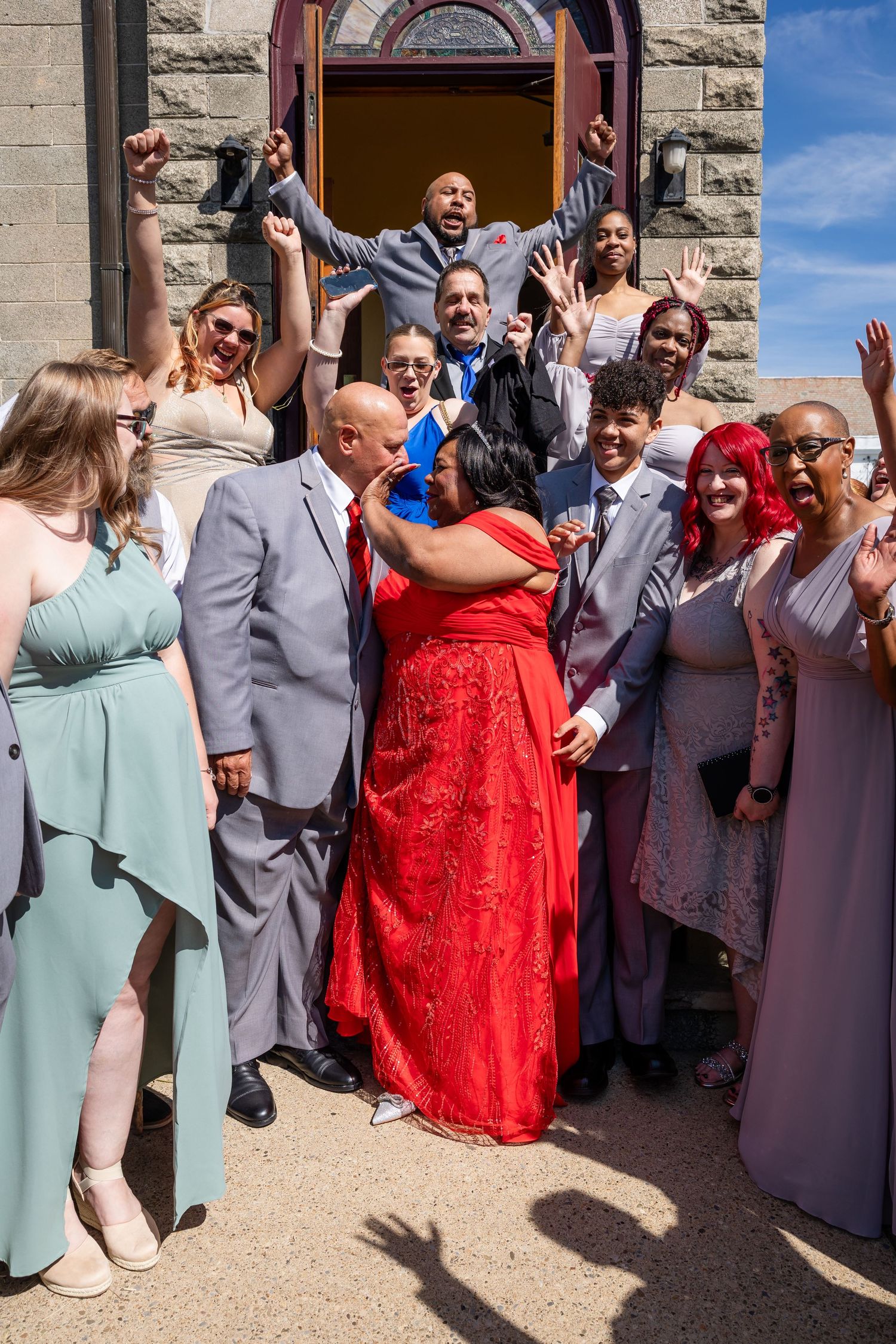 Wedding guests in colorful dresses and suits cheer with raised fists around a woman in a red gown outside a stone church.