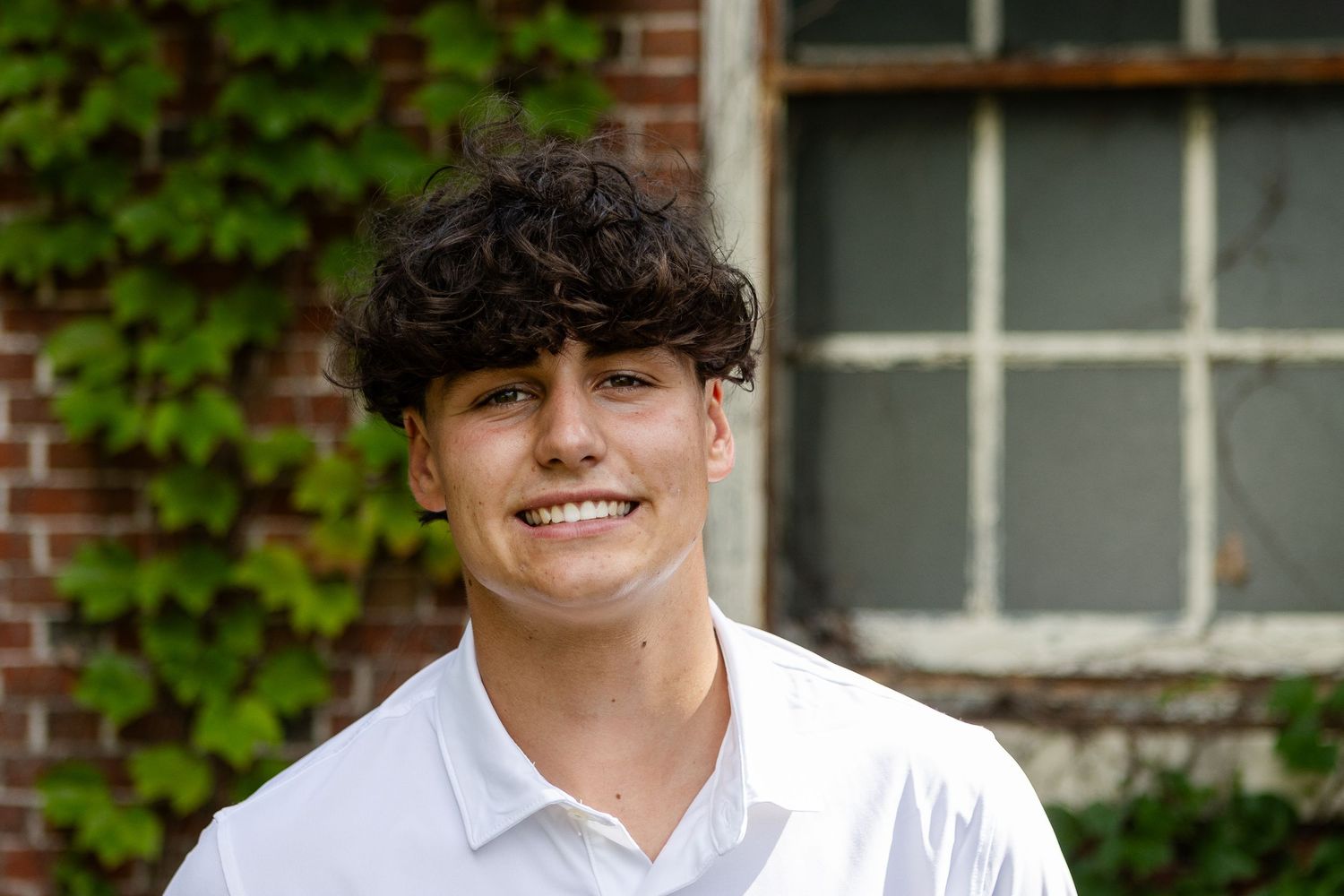 Close-up portrait of a smiling young man with dark curly hair in a white polo beside an ivy-covered brick wall.