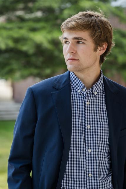 Young man in a navy blazer and checkered shirt gazes to the side with green trees softly blurred behind him.