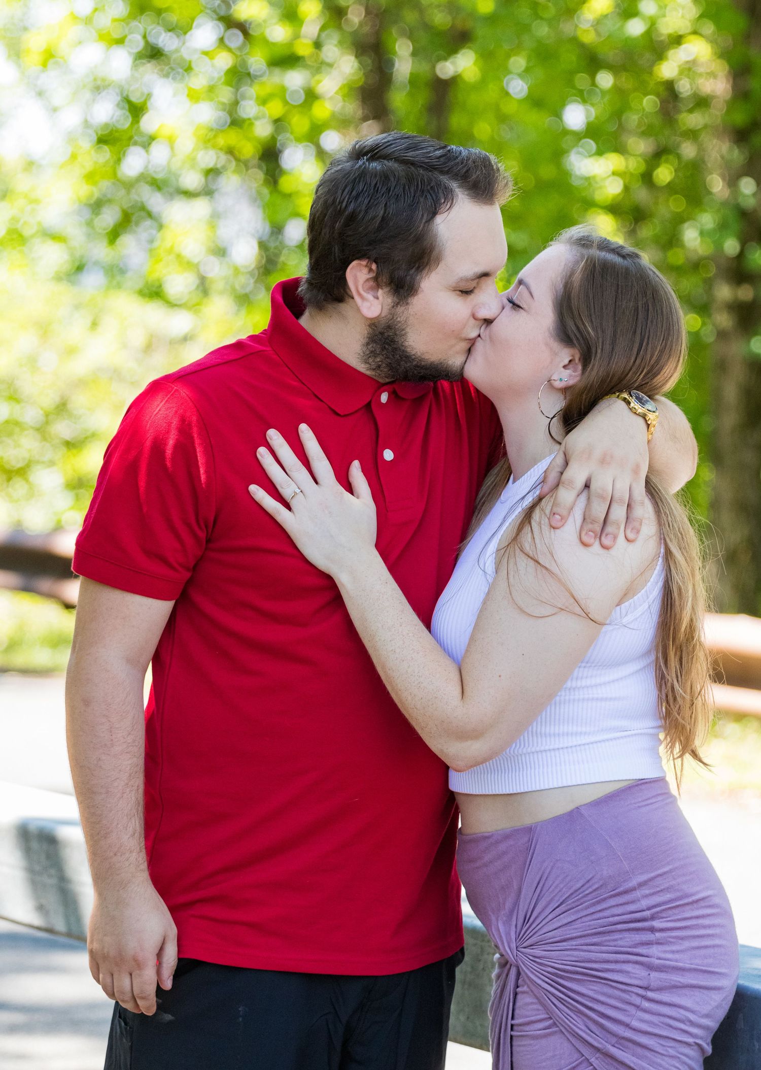 A bearded man in a red polo kisses a woman in a lavender skirt as she shows her engagement ring in a leafy park.