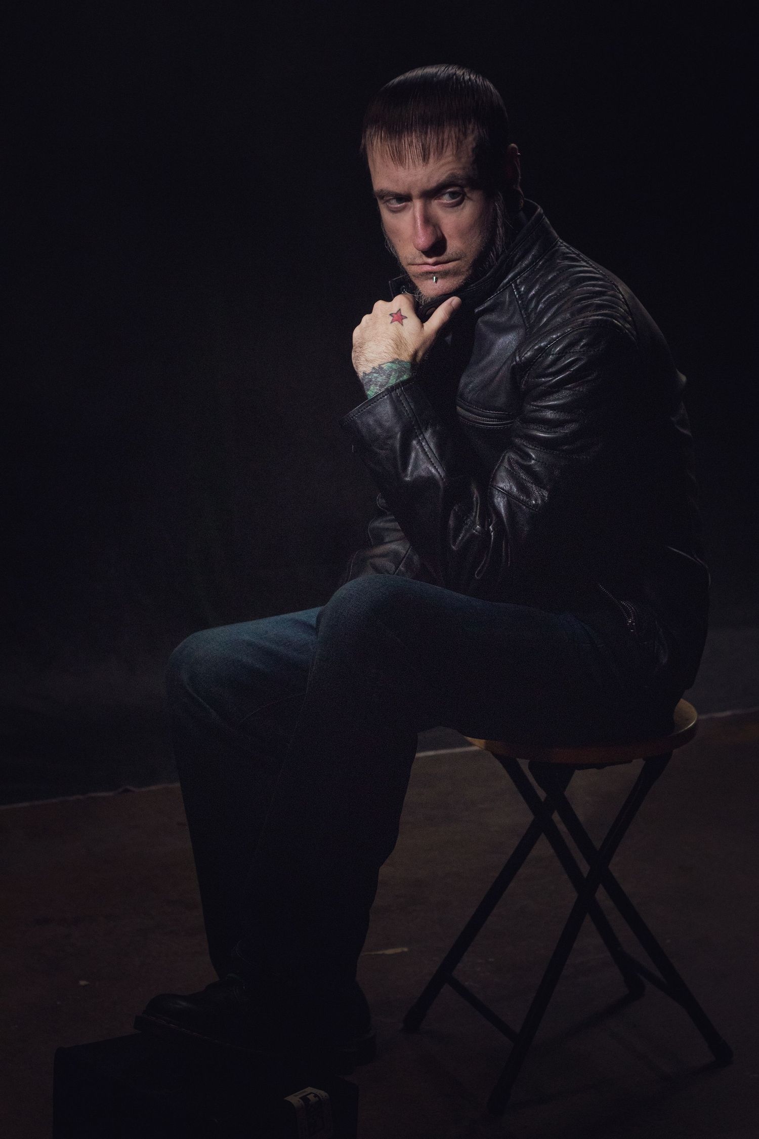 Moody studio portrait of a man in a black leather jacket sitting on a stool with his hand on his chin against a dark background.