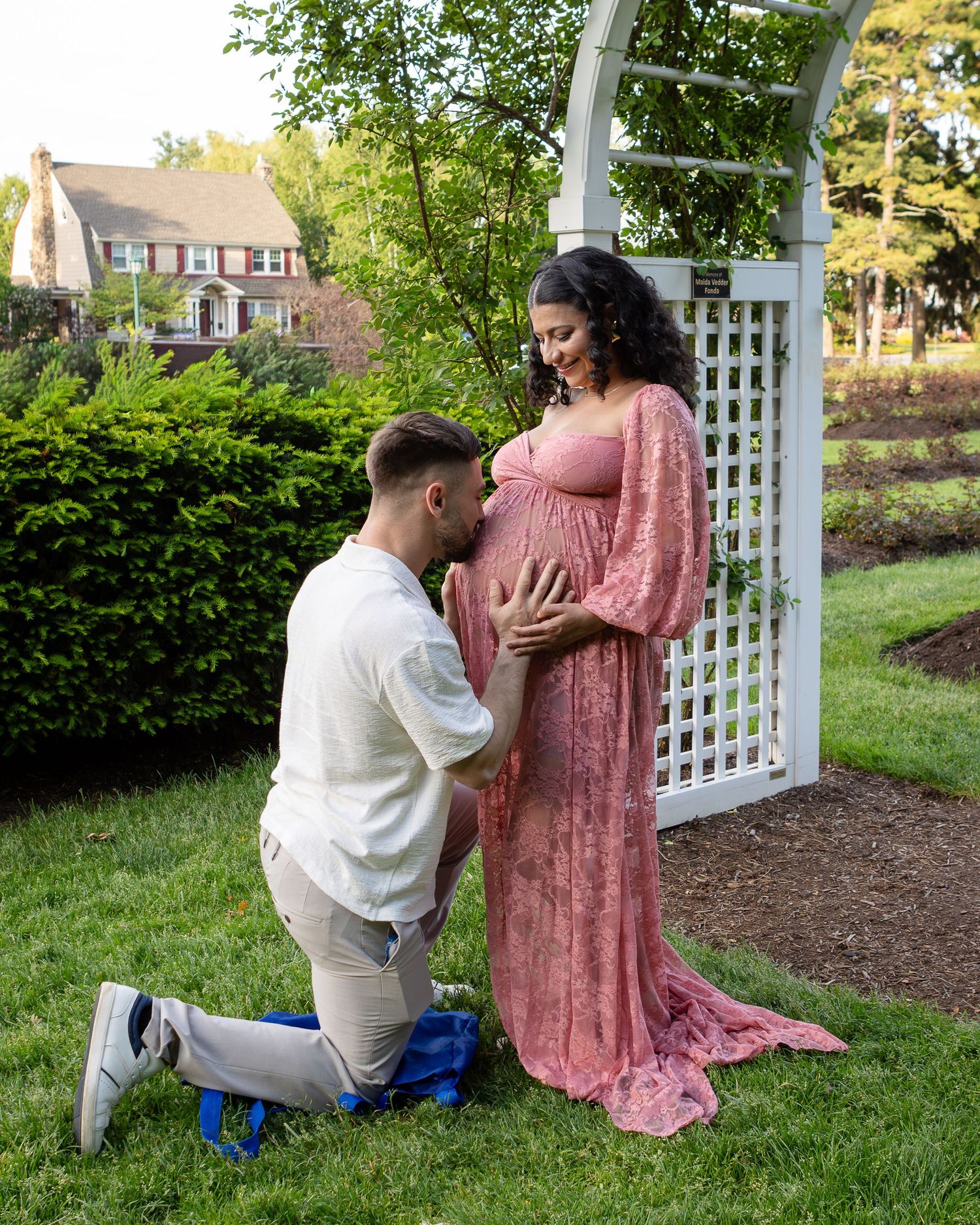 A man kneels on the grass to kiss his pregnant partner's belly beside a white garden arbor.
