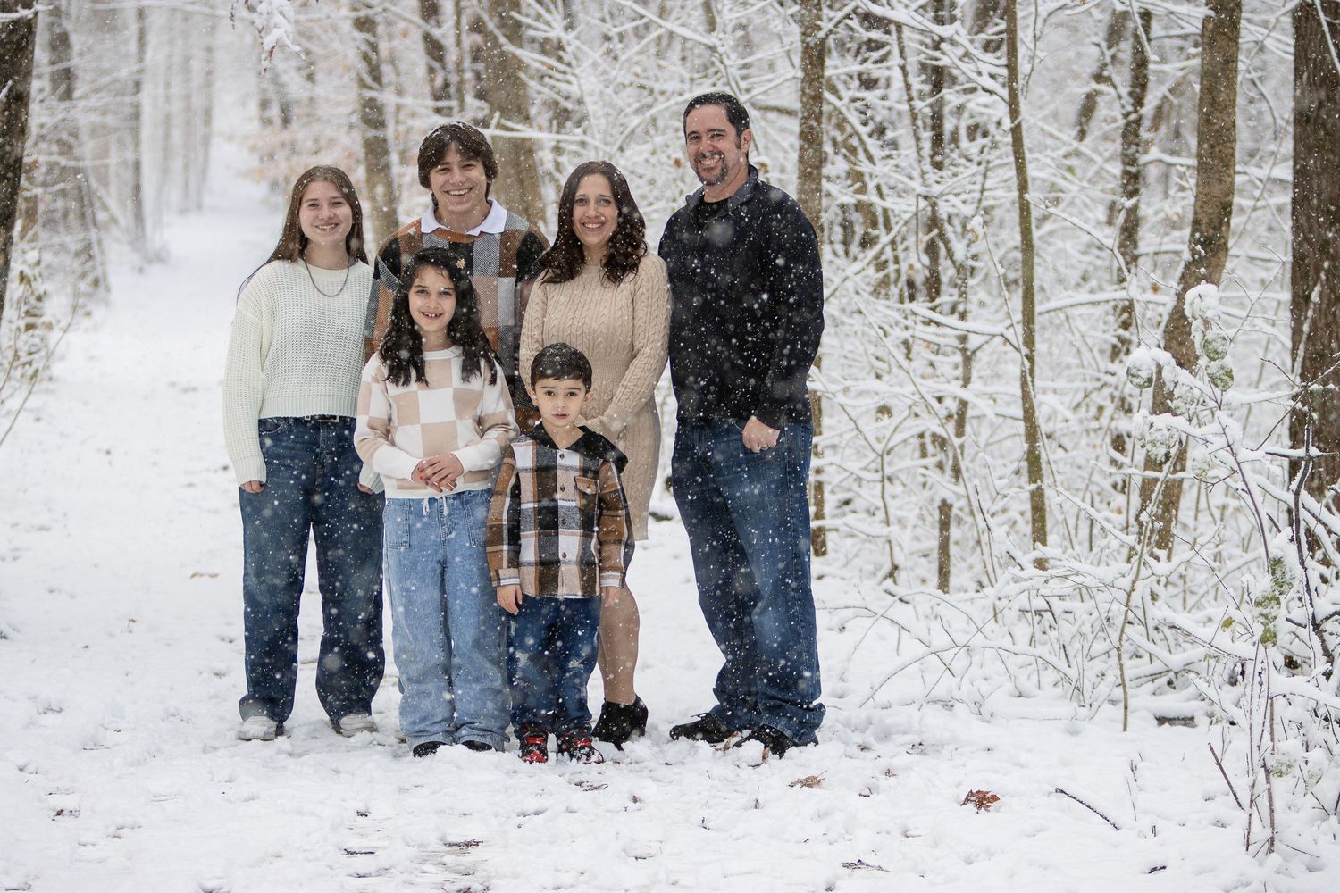 Family of six in warm casual clothes poses together on a snowy forest trail with frosted trees in the background.