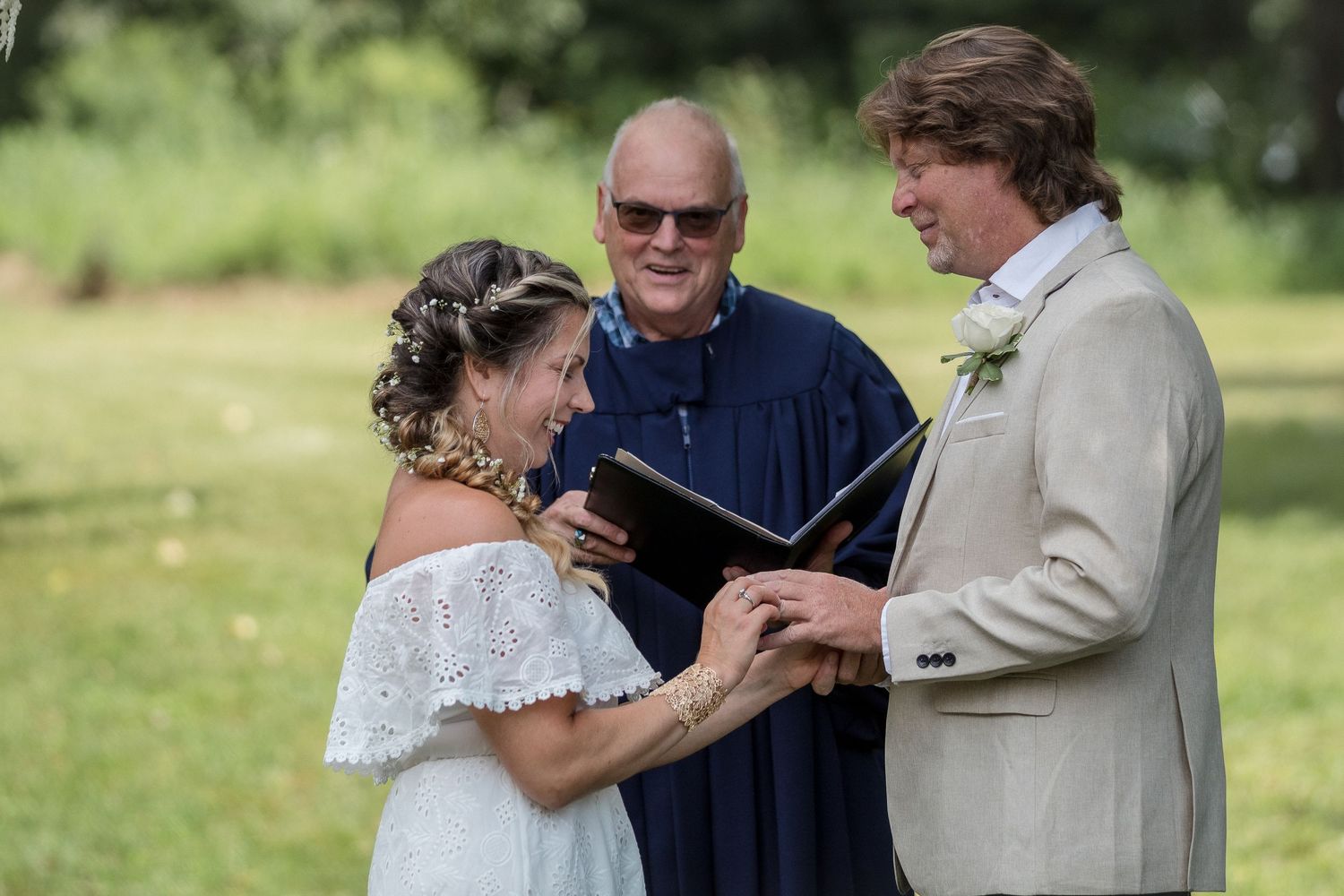 A bride and groom hold hands during an outdoor wedding ceremony while a smiling officiant in a blue robe reads from a book.