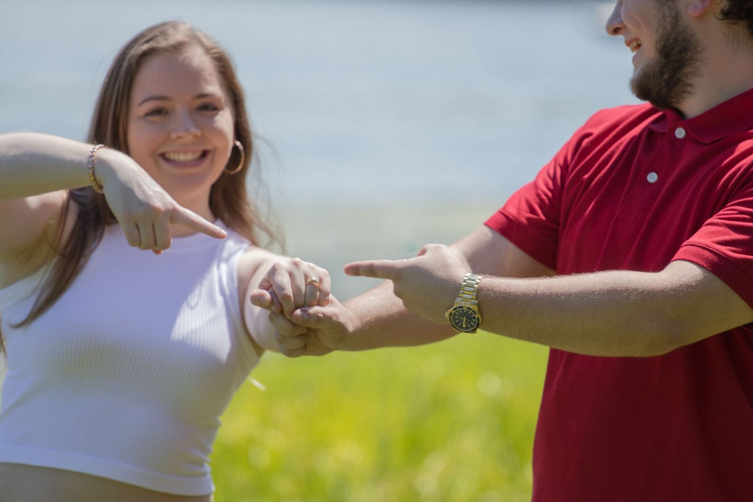 A smiling woman and a man in a red polo hold hands and point at her engagement ring near a sunny lake.