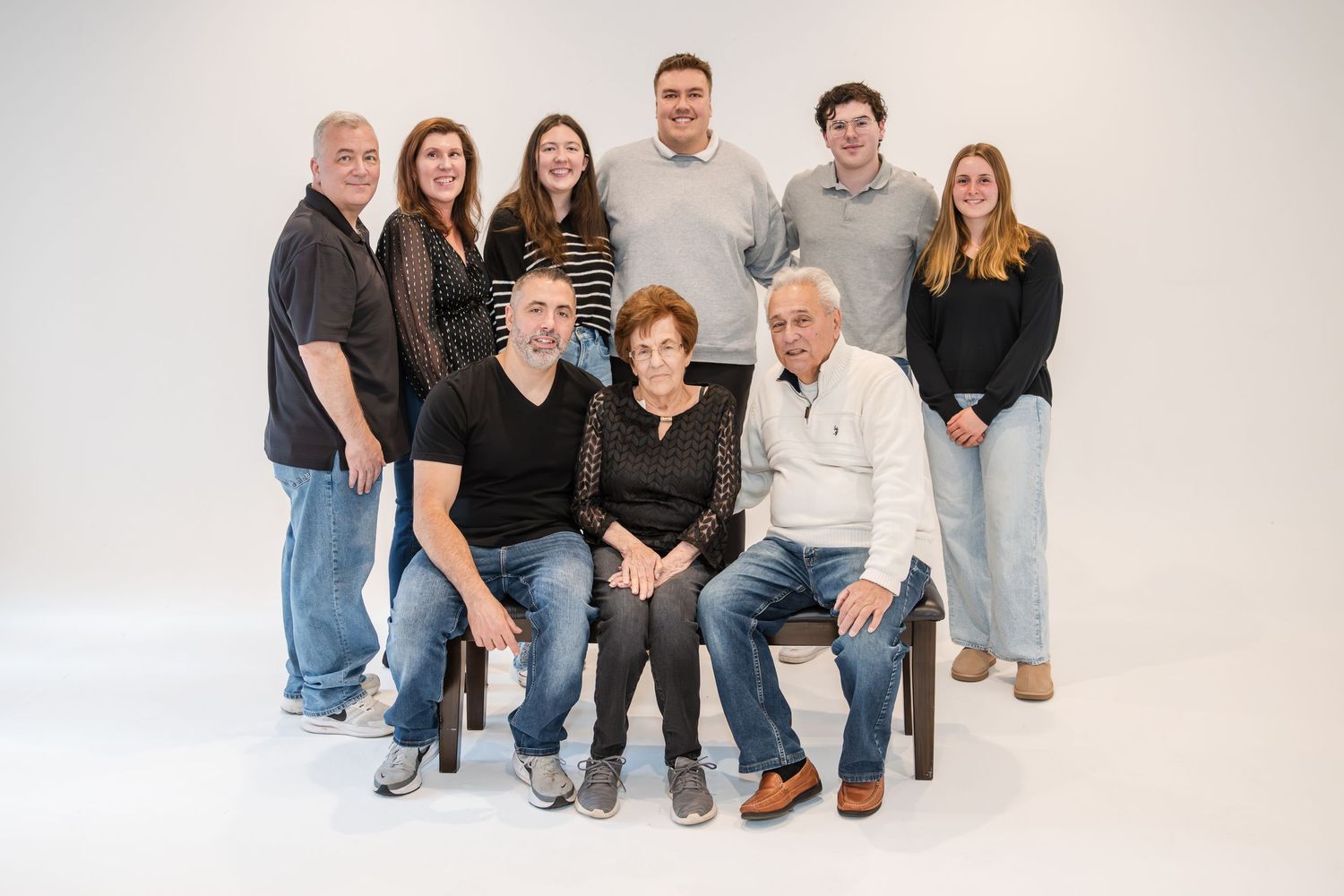 A multigenerational family of nine poses together in a bright studio, with grandparents seated in front.