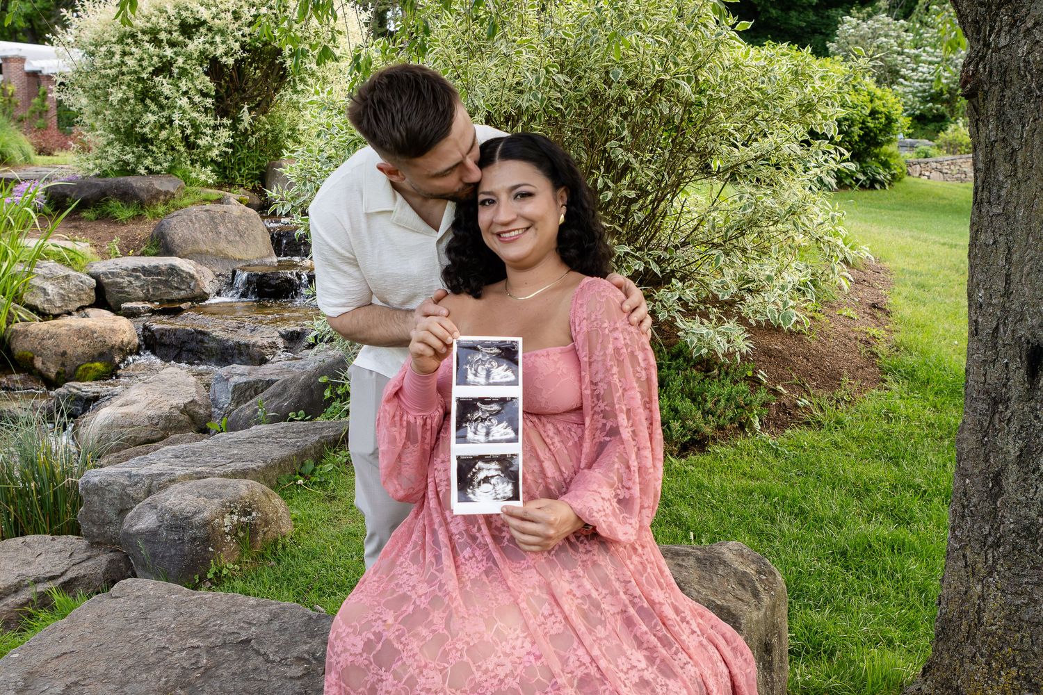 A smiling pregnant woman holds ultrasound photos as her partner kisses her cheek beside a rocky stream.