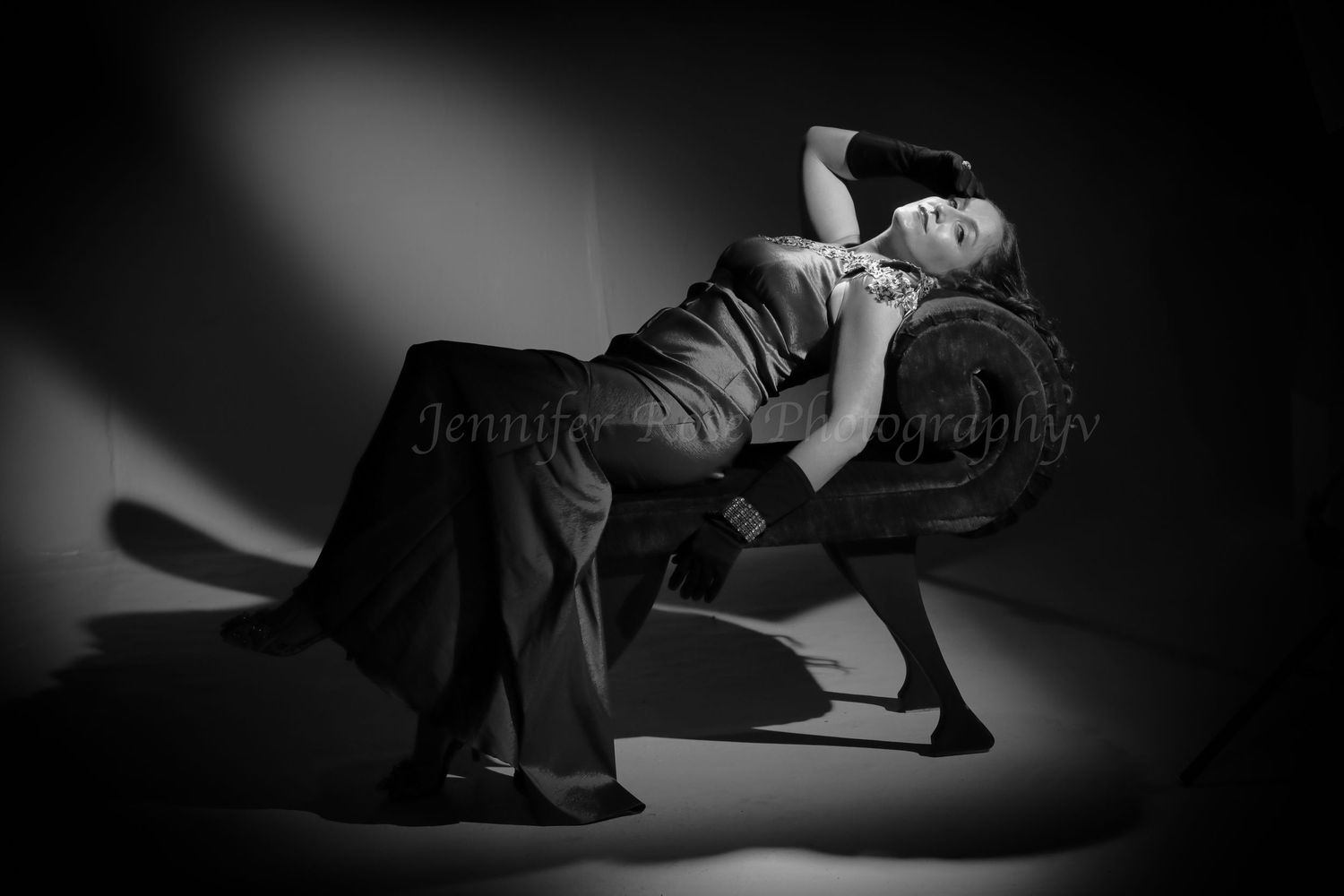 Black and white film noir portrait of a woman in an evening gown reclining on an antique chair with dramatic spotlight shadows.