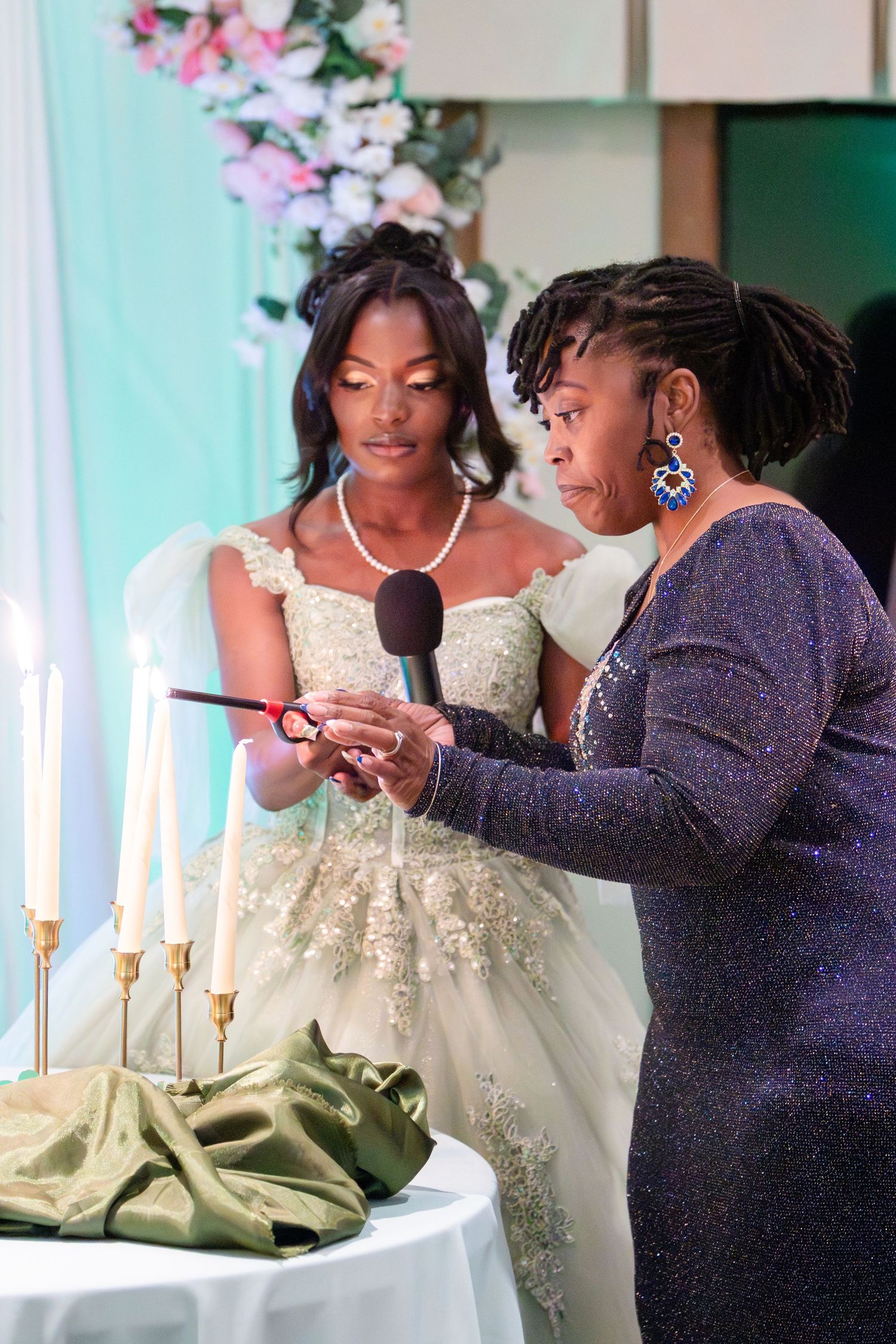 A bride in a beaded ivory gown and a woman in a sparkly dress light candles together during a wedding unity ceremony.