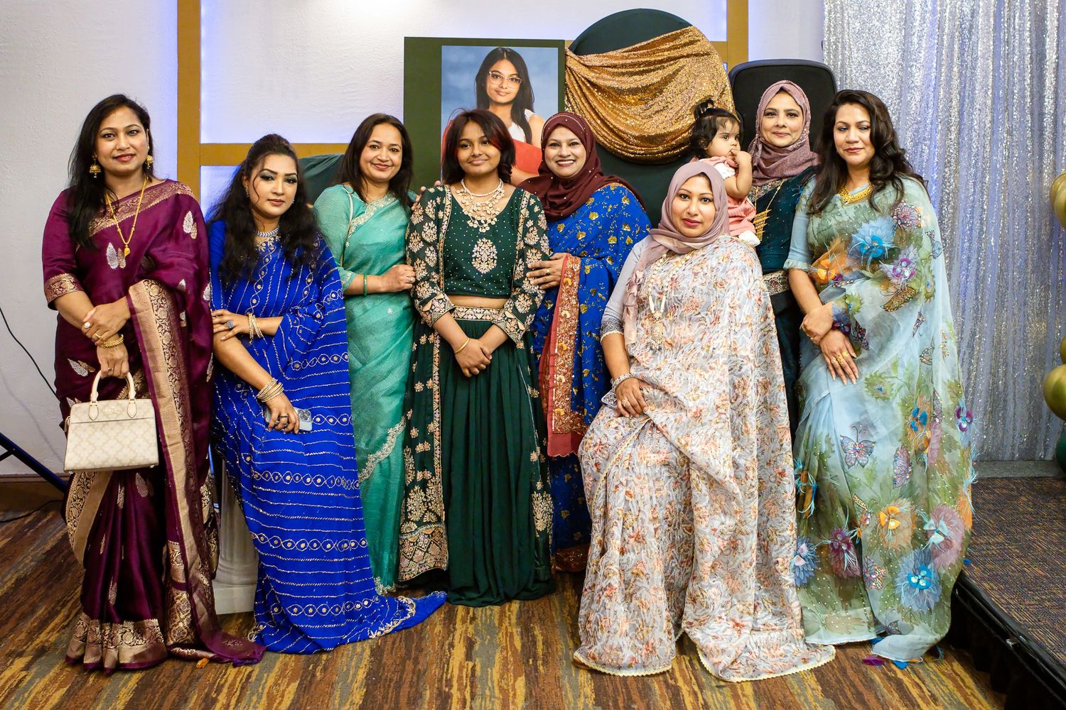Group of smiling women in colorful silk sarees and traditional South Asian dresses posing together on a stage at an elegant indoor event.